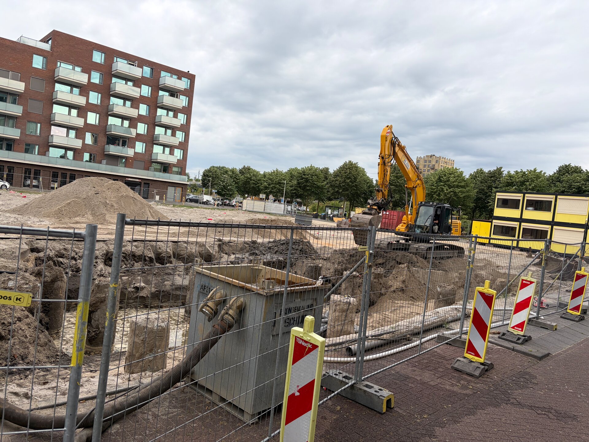 Excavator removing soil between foundation piles, open pit visible from the street