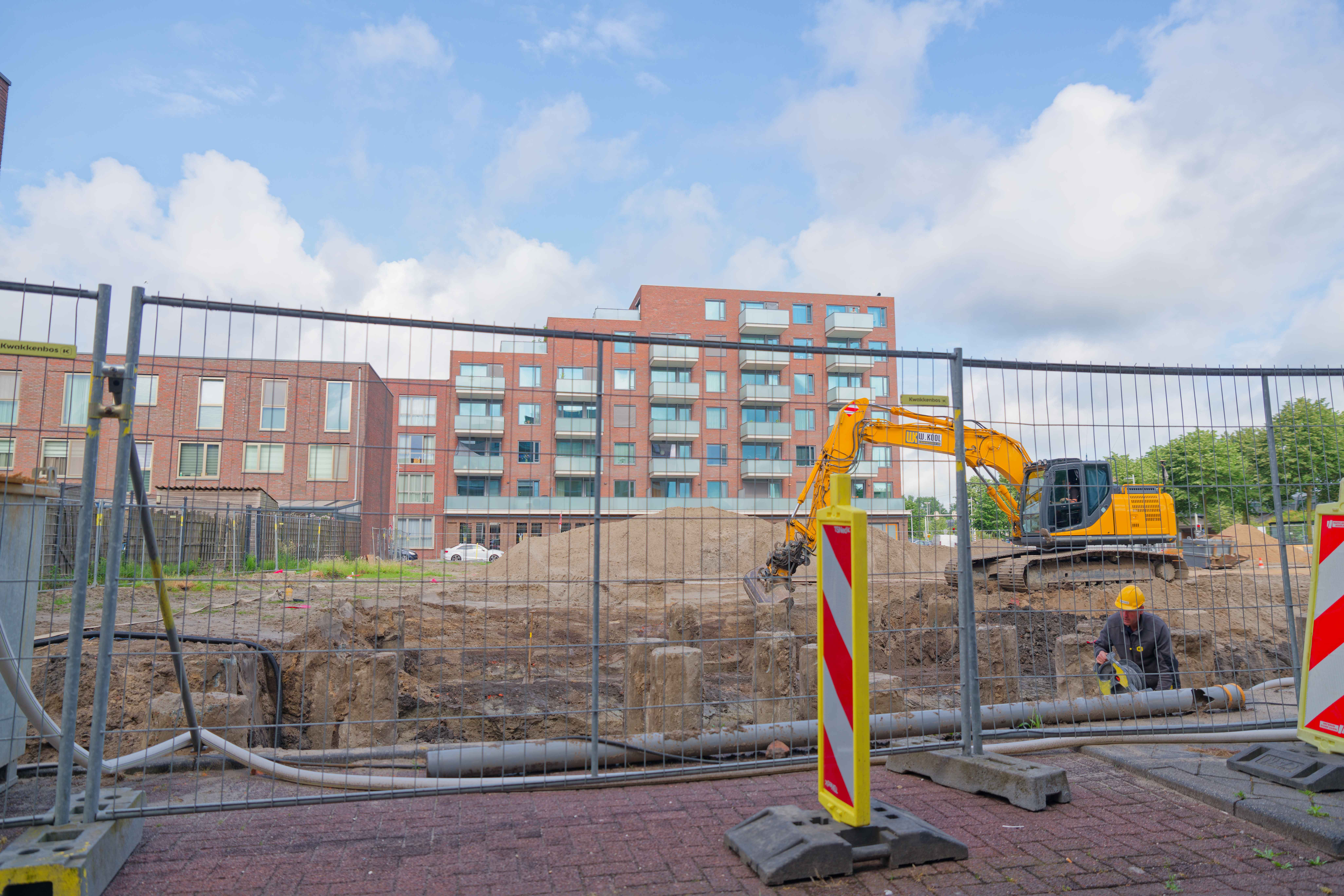 JCB excavator digging the pit between foundation piles, worker in hard hat visible, viewed through security fence