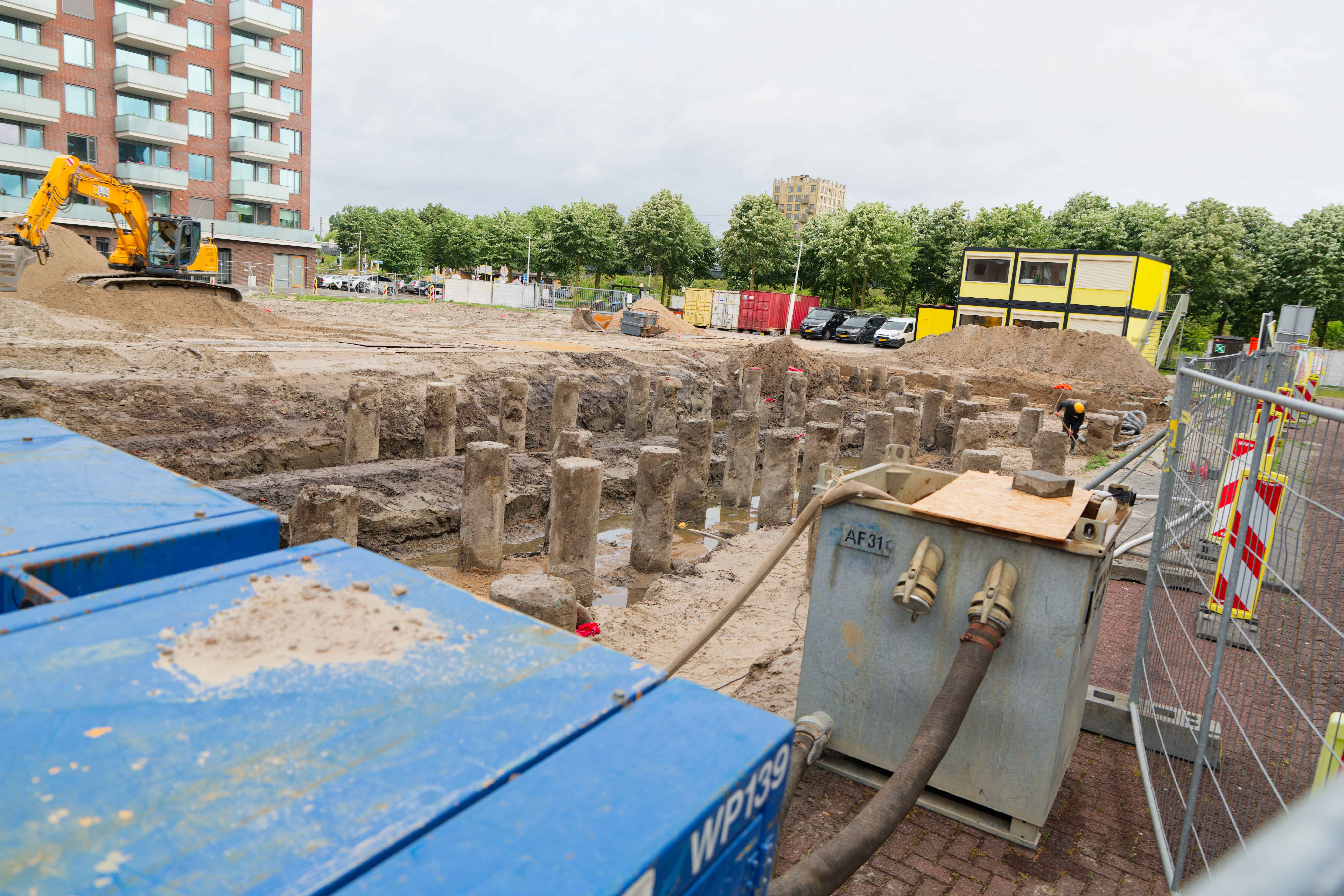 Rows of exposed concrete foundation piles in the open pit, excavator and apartment building behind, seen over the security fence
