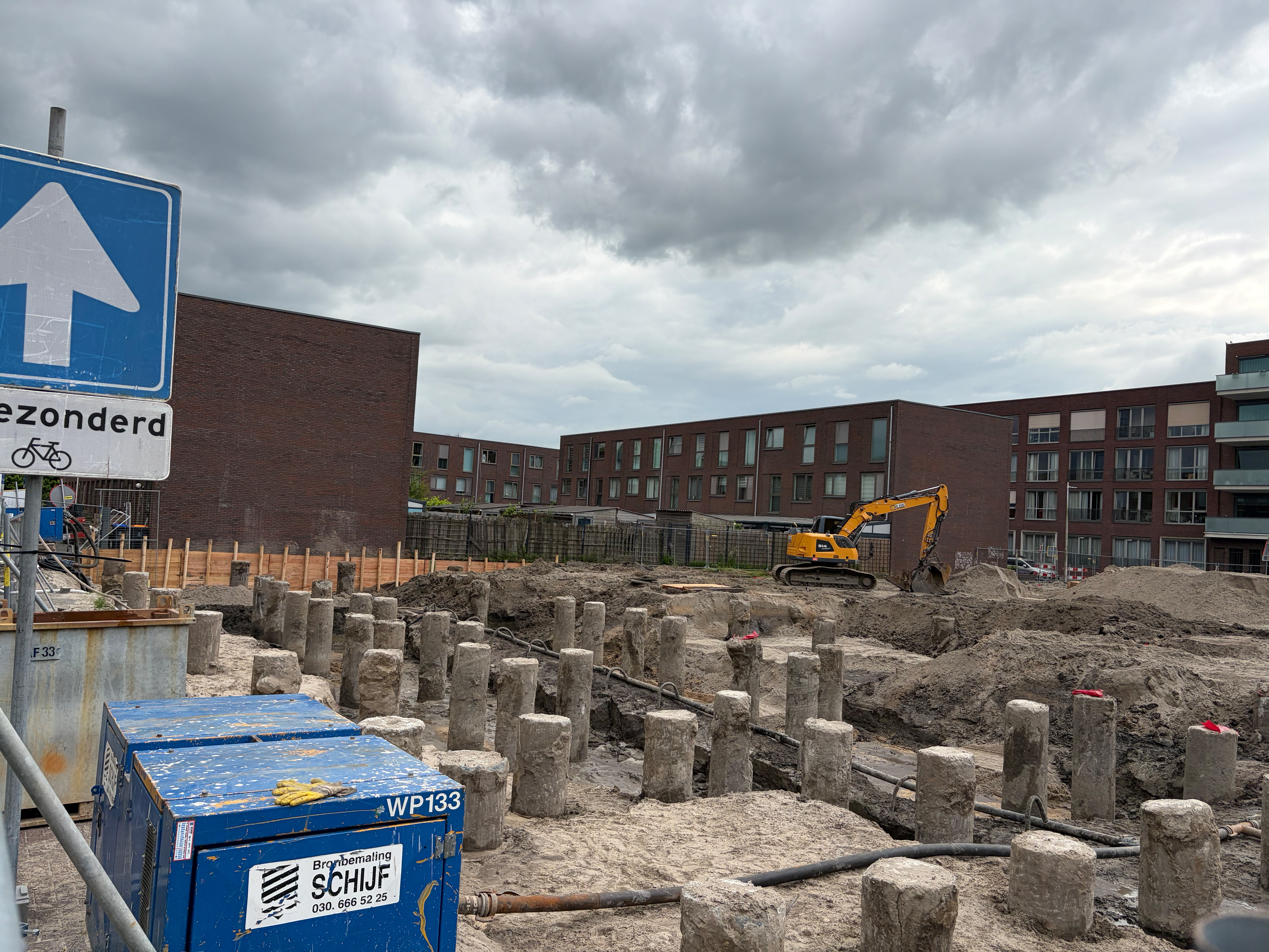 Wide view of the foundation-pile field with a JCB excavator and the apartment building behind, a blue street sign in the foreground