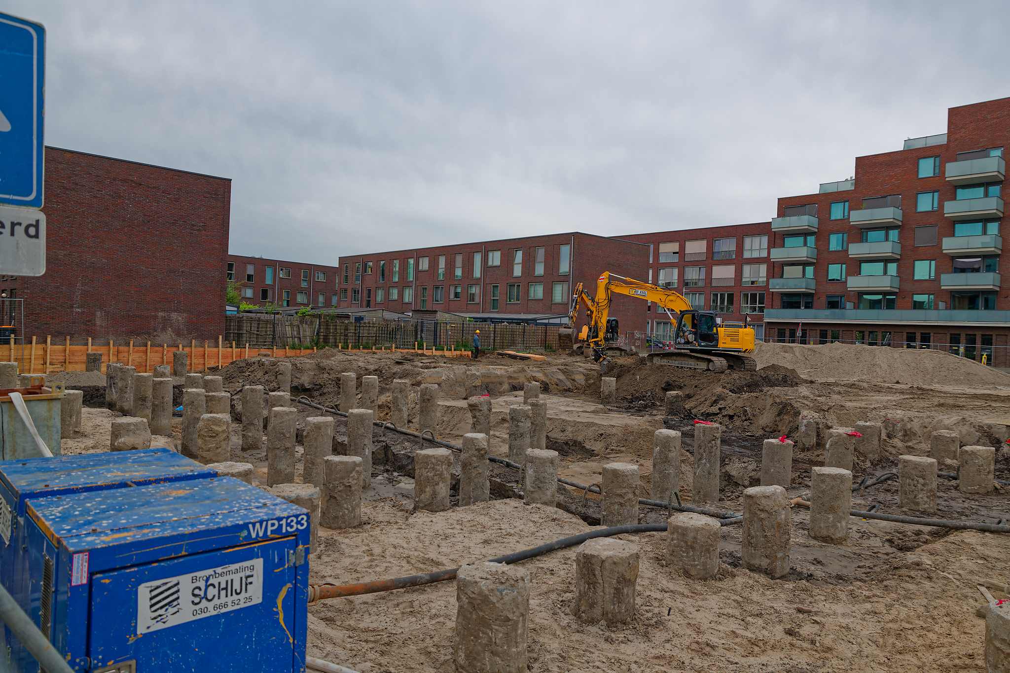 Field of exposed concrete foundation piles in the sandy pit, a yellow excavator at work with apartment buildings behind, seen over the security fence