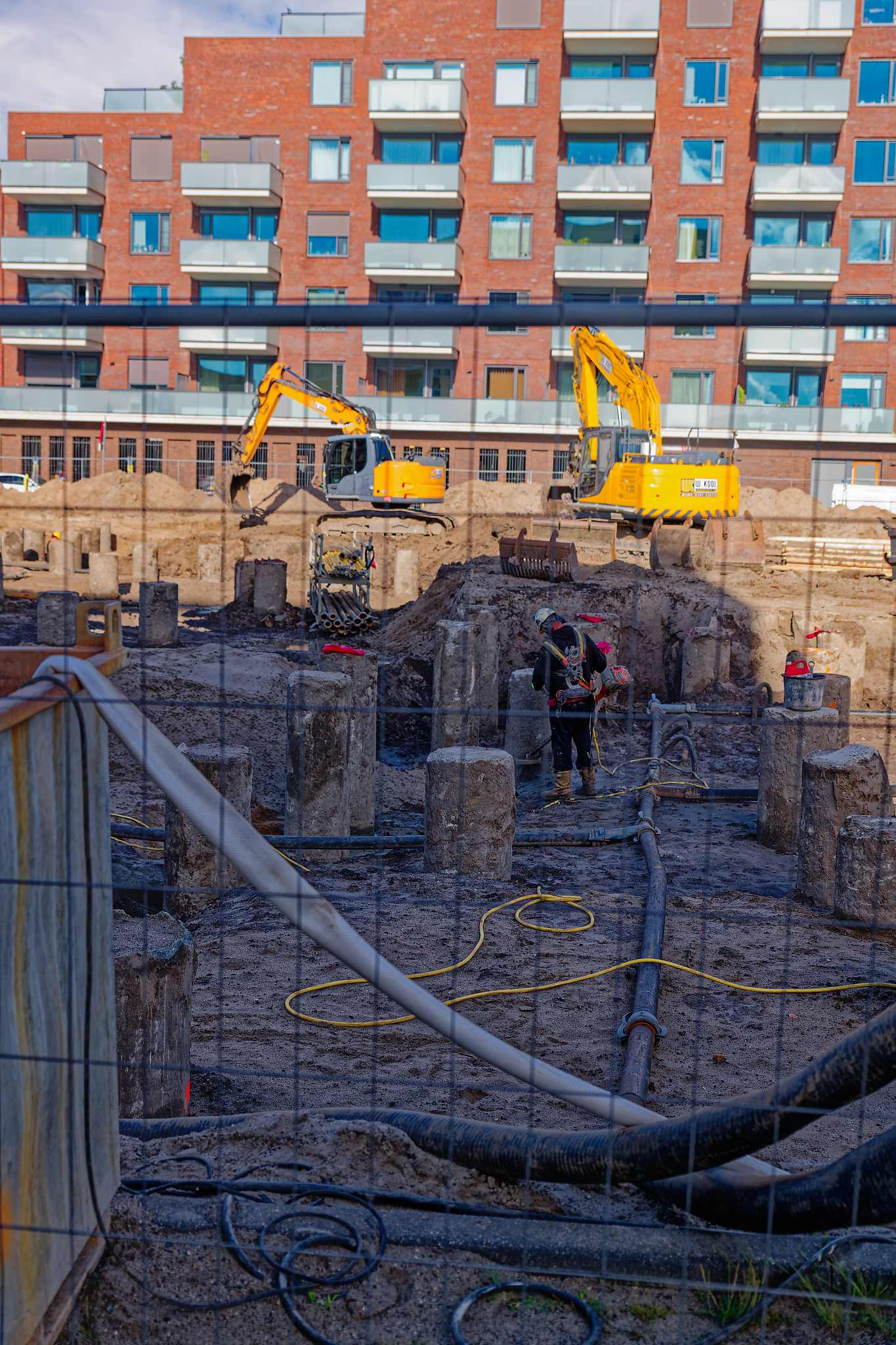 Two yellow excavators and a worker among the concrete pile heads, dewatering hoses crossing the pit, seen through the security fence