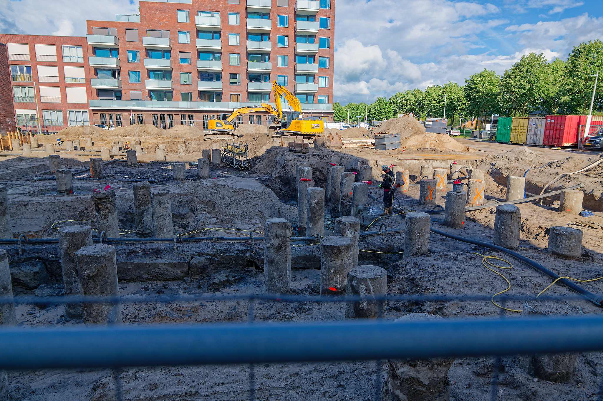 Wide view of the pit full of cropped pile heads, an excavator on the sand and a blue pipe crossing the foreground