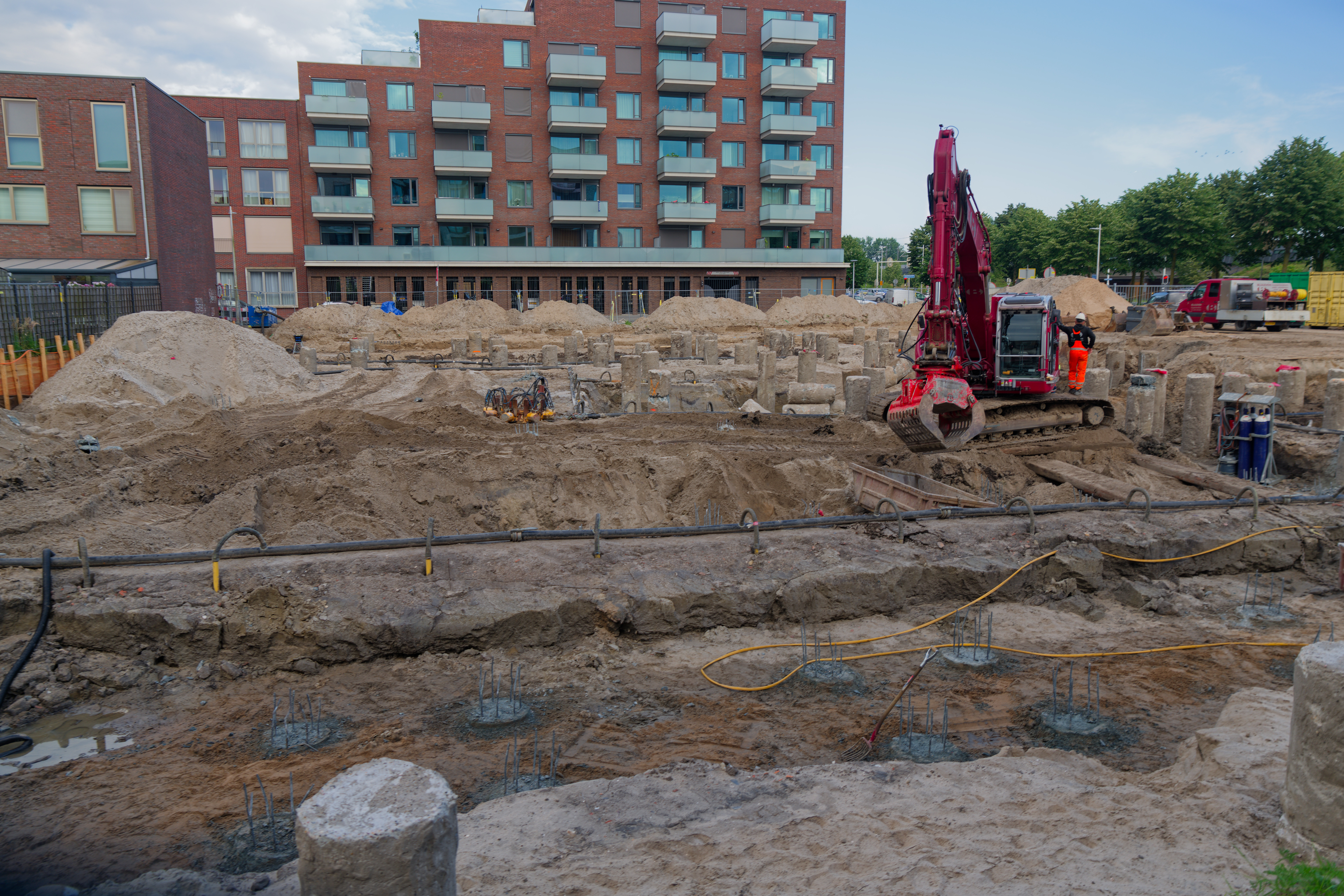 A red tracked excavator works the sandy foundation pit, cropped concrete pile heads with protruding rebar in the foreground, the brick Circulus apartments behind, seen over the security fence