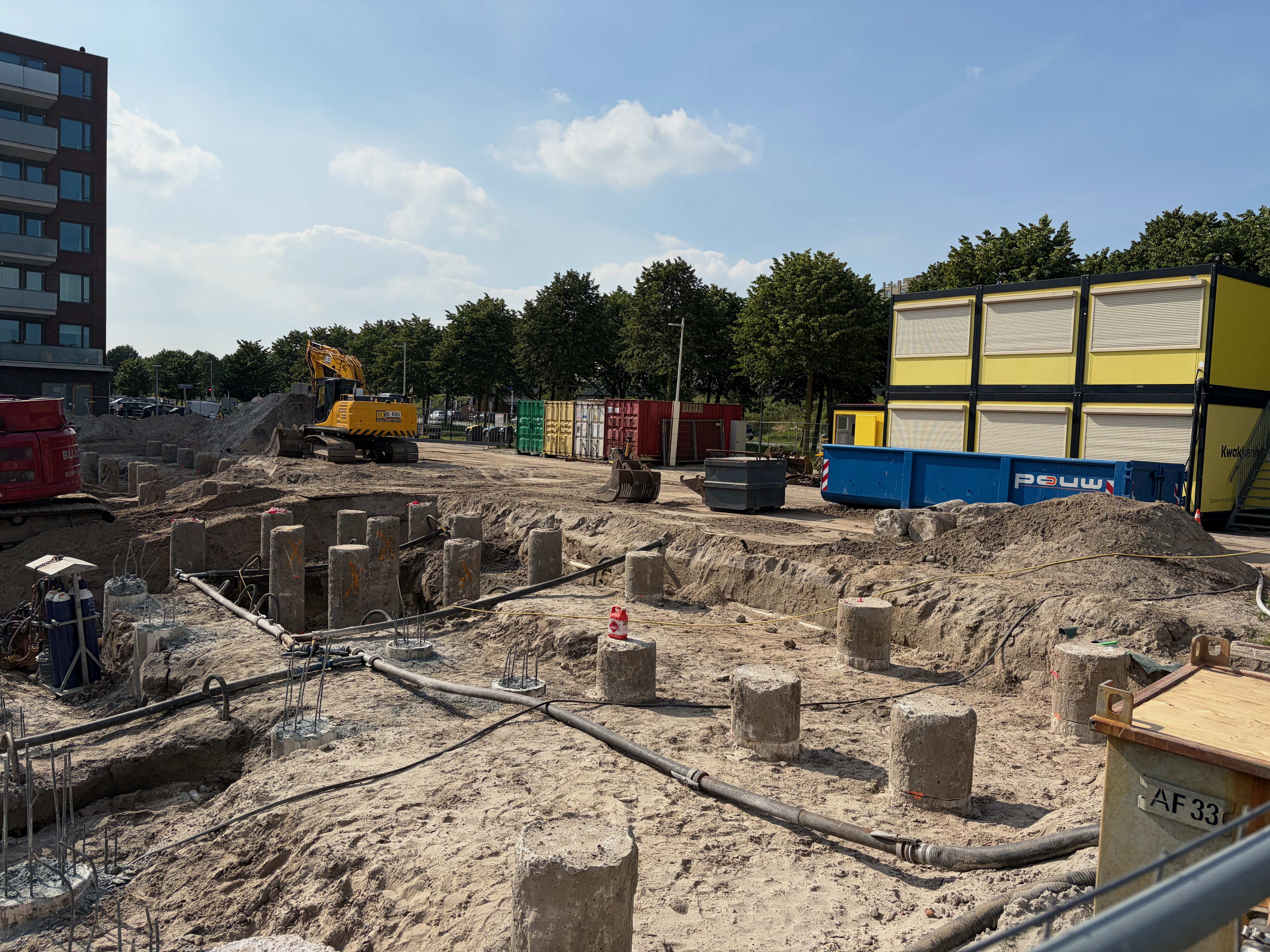 Wide view of the sandy pit with rows of cropped concrete pile stumps tipped with rebar, a yellow excavator, shipping containers and a skip along the far edge under a blue sky