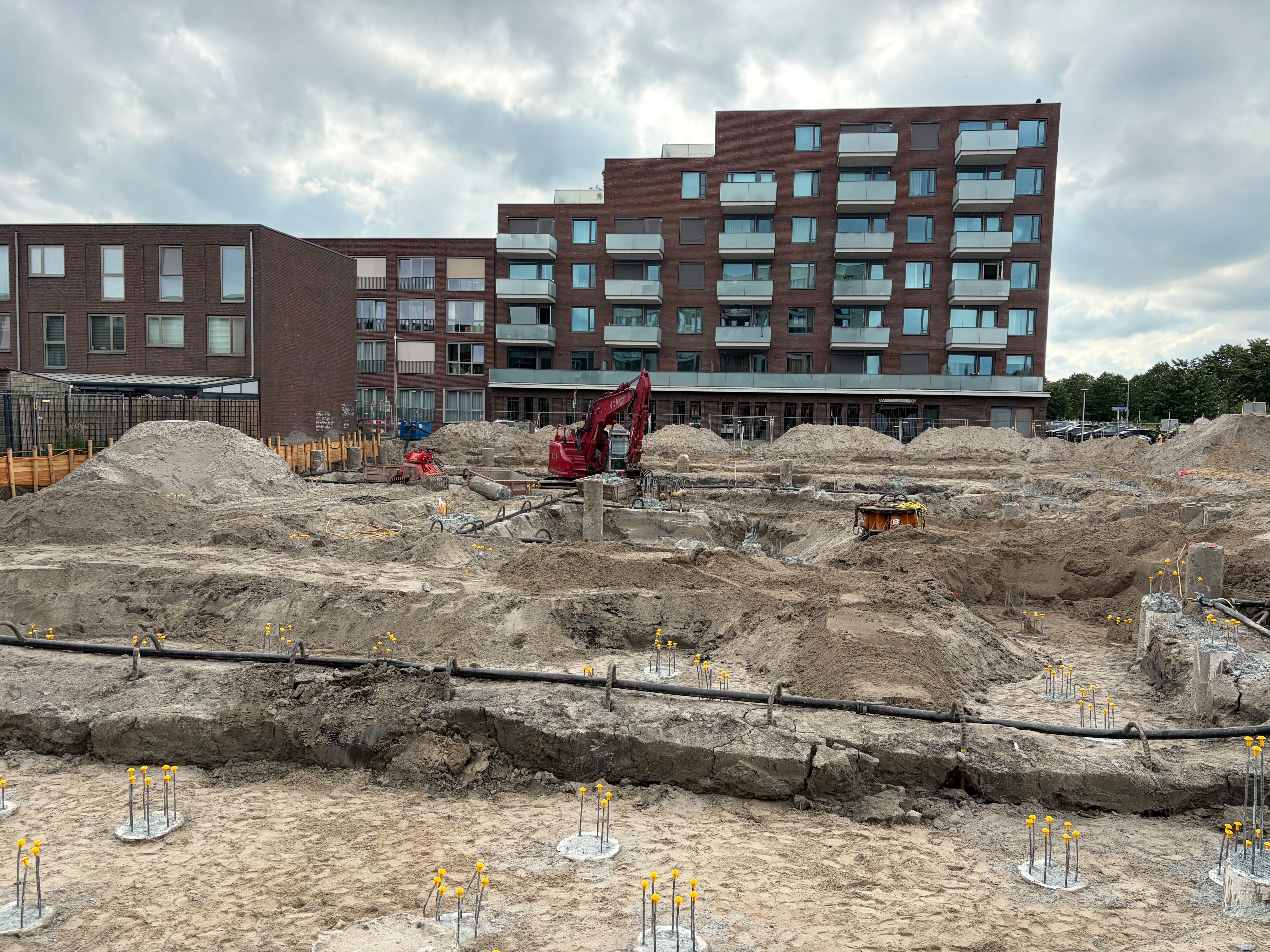 The foundation pit under a grey sky, rows of slender rebar cages capped with yellow safety cups in the foreground sand, a red excavator mid-pit and black dewatering hoses looping across the floor, the brick Circulus apartments behind