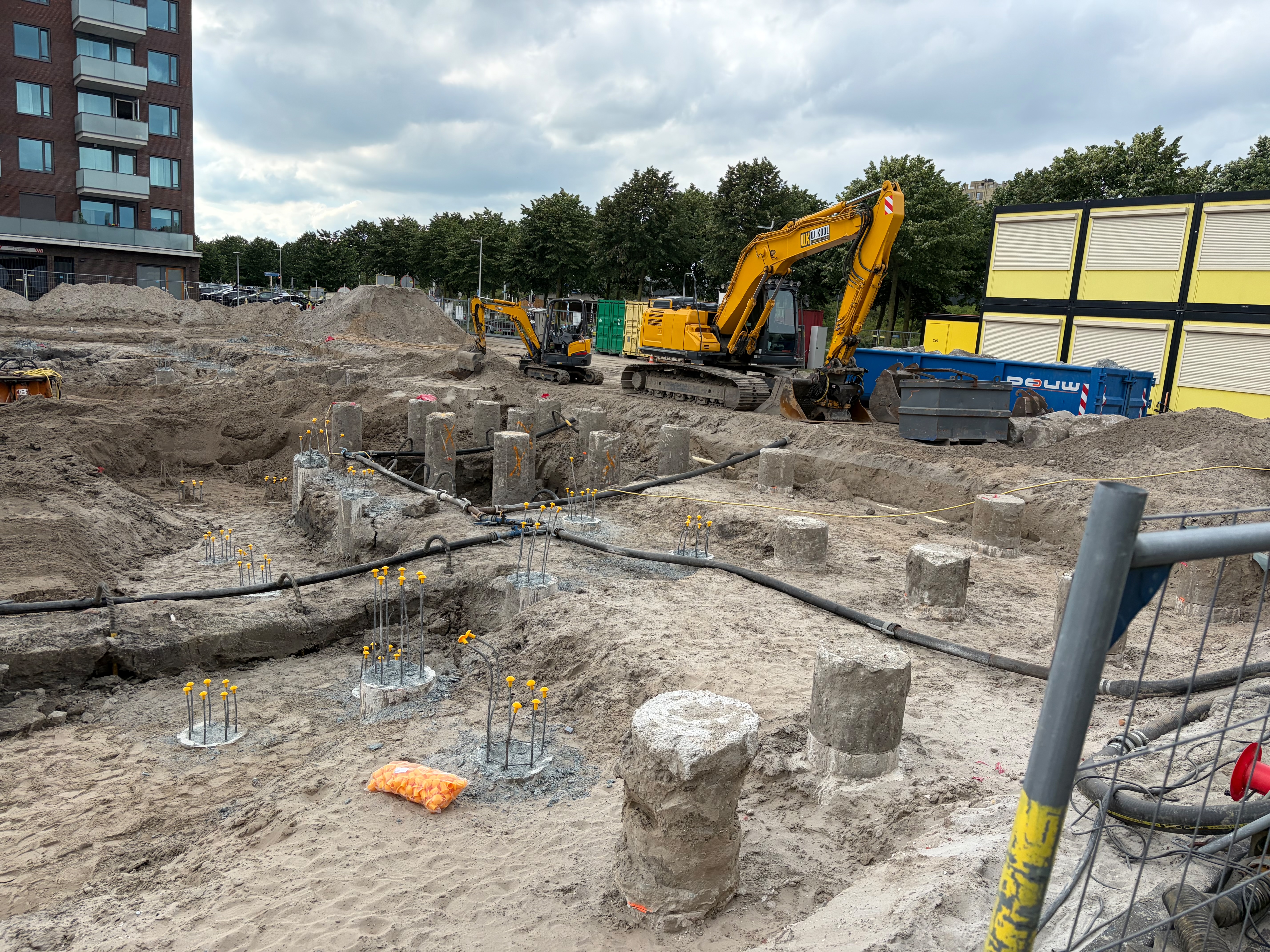 Close view of thick round grey concrete pile stumps with rebar bursting from the top, a yellow CAT excavator and a smaller machine working the back of the site beside shipping containers, a blue skip and yellow hoarding panels