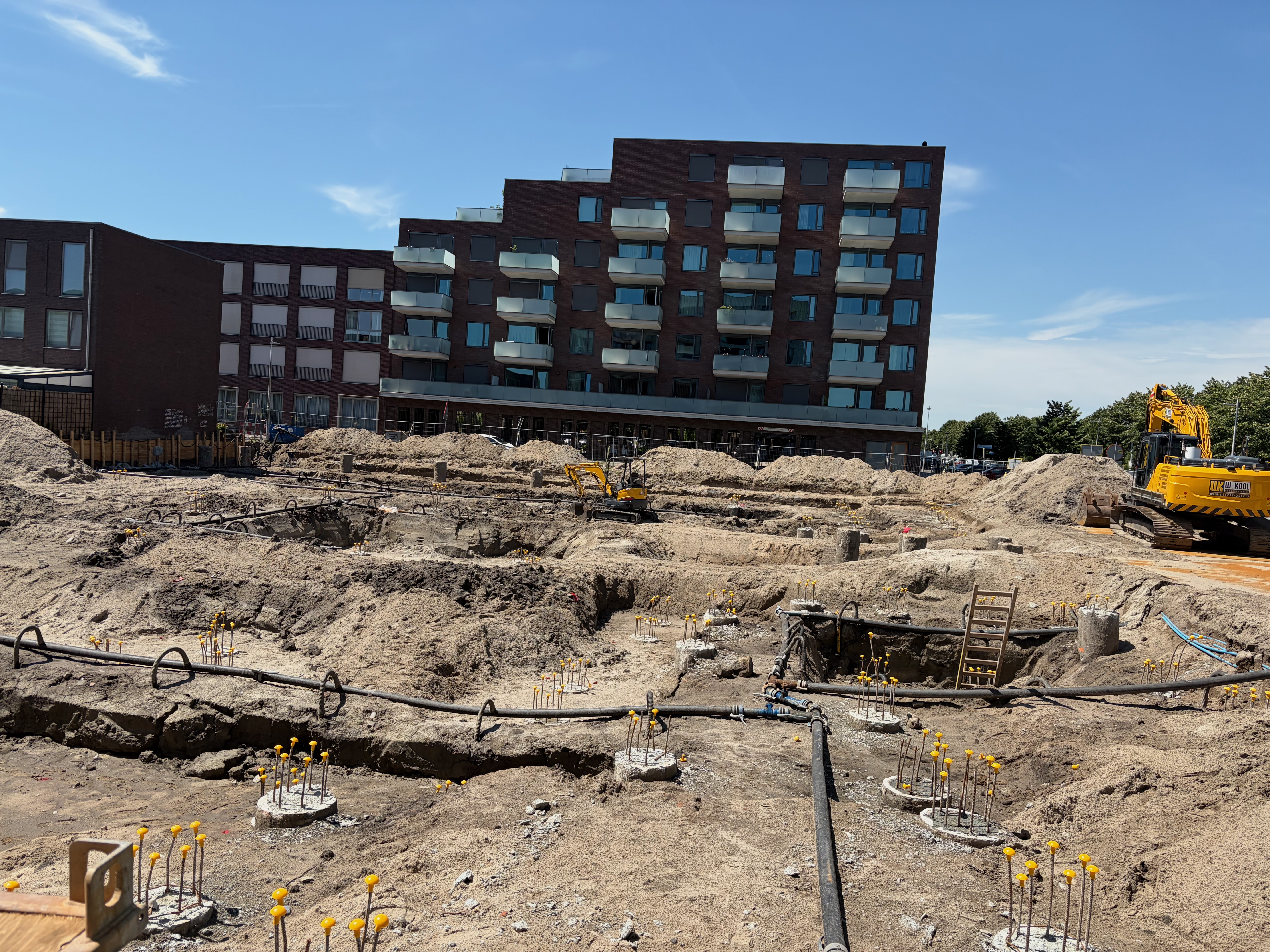 Wide sunny view of the entire foundation pit under a deep blue sky, dozens of rebar cages tipped with yellow safety caps arranged in a grid from foreground to the brick Circulus apartment block, two small excavators working mid-pit and dewatering hoses across the floor