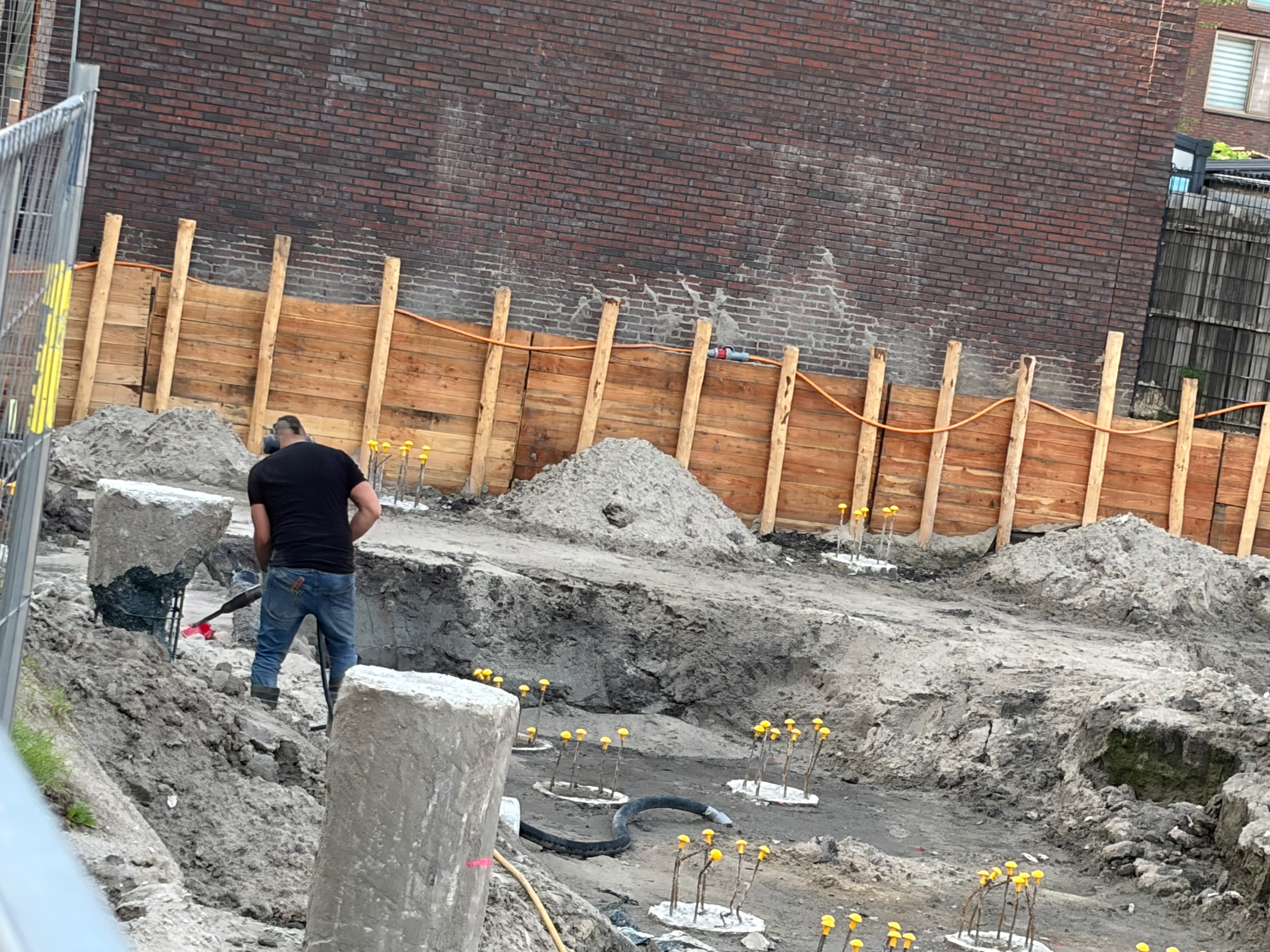 Worker in black t-shirt shovelling sand by hand around concrete pile stumps, yellow-capped rebar cages visible in the middle distance, wooden hoarding fence and orange safety netting behind