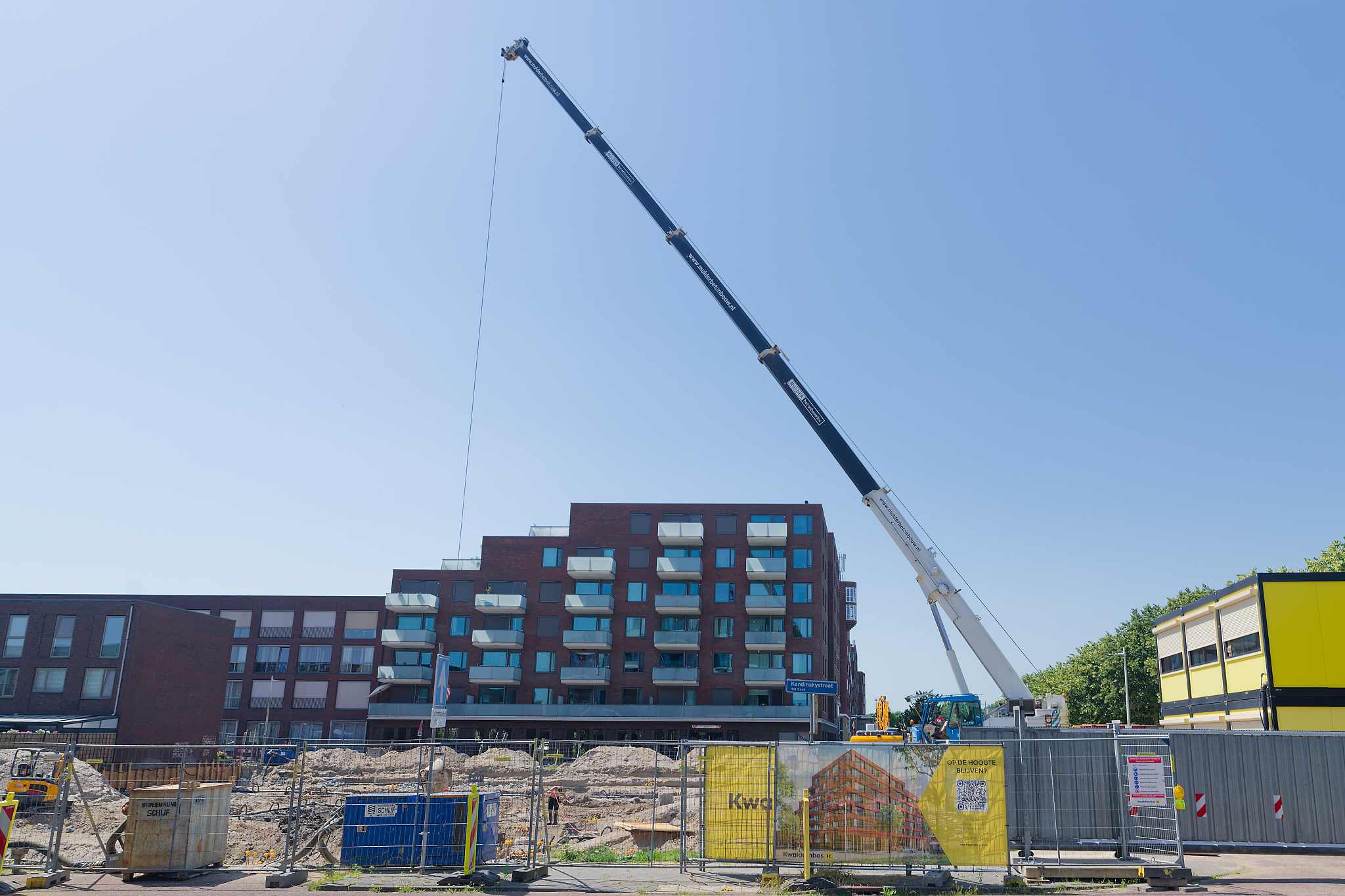 Wide street-level view of the mobile crane boom reaching diagonally across the blue sky, the Circulus apartment blocks behind, construction site with sand mounds and workers below
