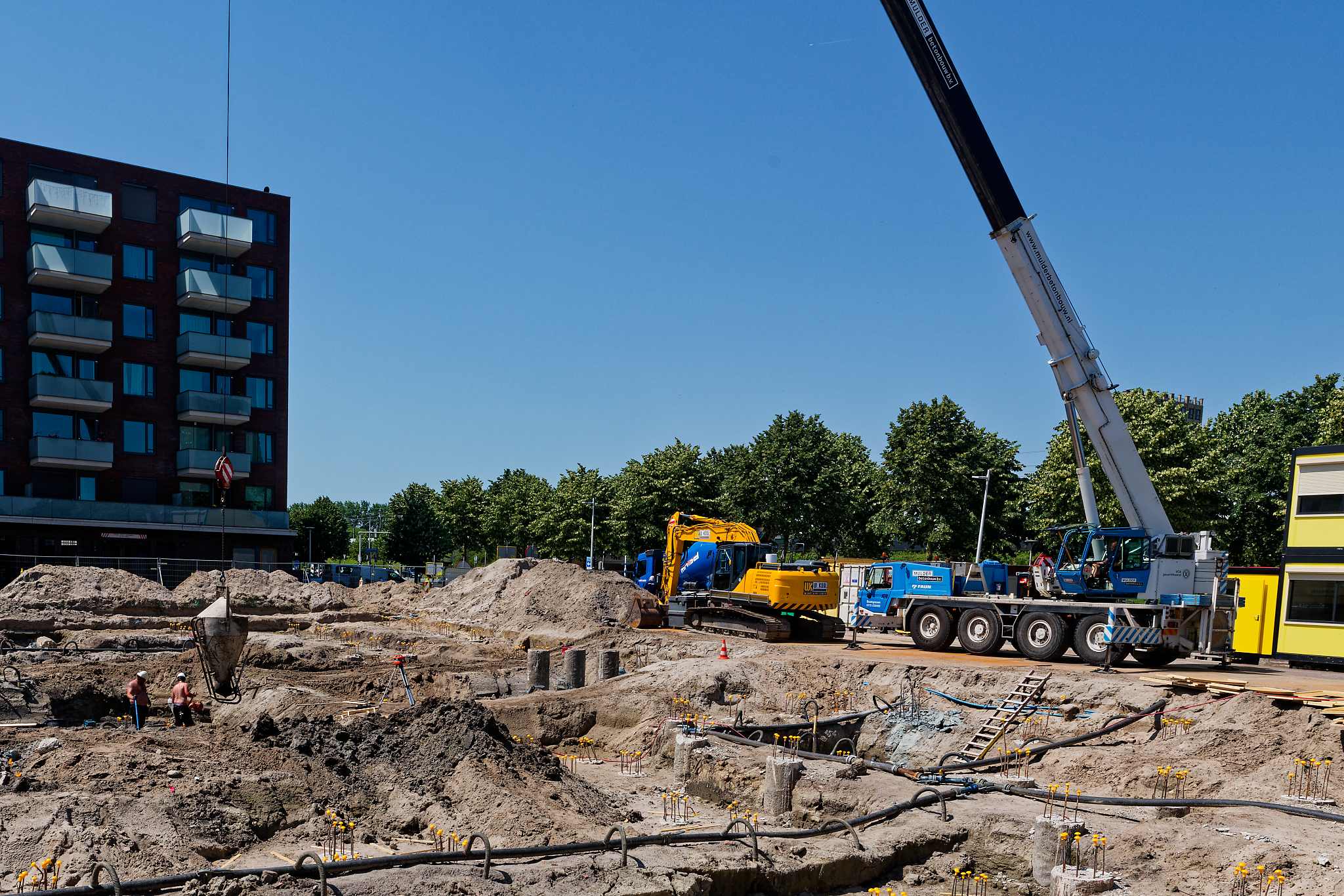 Overview of the full construction site from street level, crane visible on the right edge, yellow fencing with project sign in foreground, sandy mounds and pile work filling the entire footprint