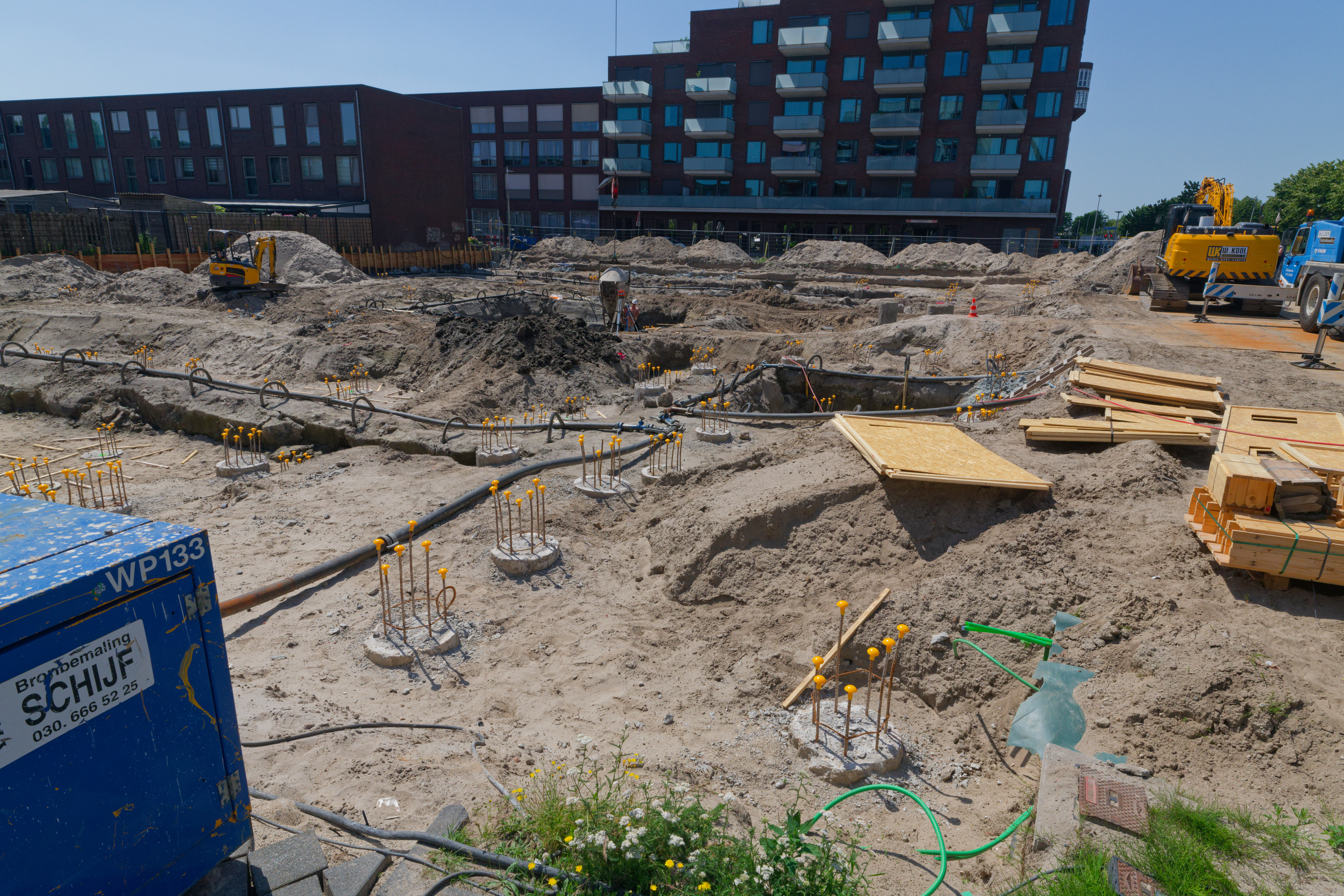 View into the foundation pit: blue dewatering pump Schijf WP 133 in the foreground, timber formwork planks stacked beside the pile caps, rebar cages throughout, excavator and crane at the far end under a blue sky