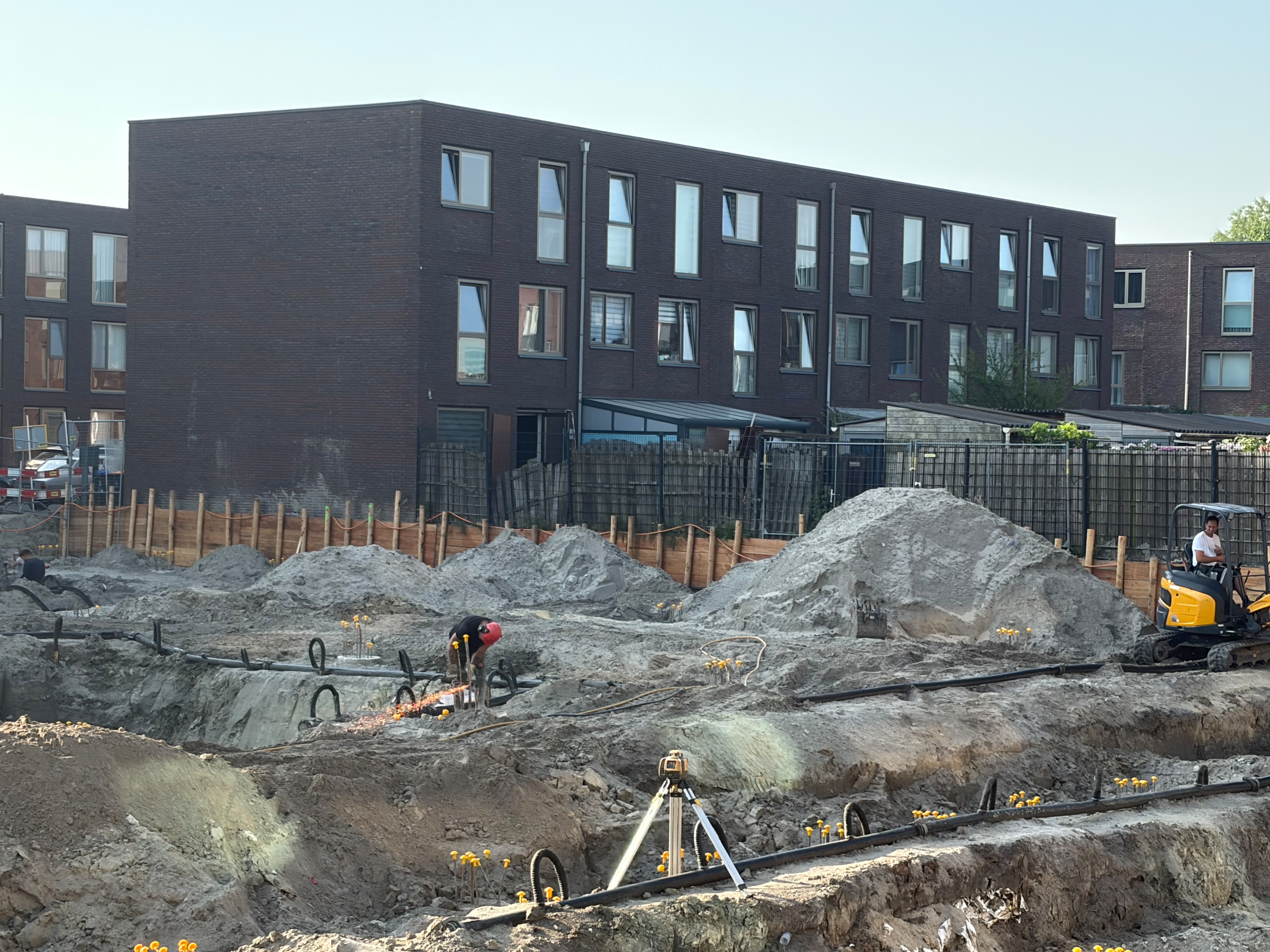 Foundation pit the day after the first pour: a total station on a tripod in the foreground right, a compact yellow excavator to the right, yellow-capped rebar stubs dotting the sandy pit floor, workers visible, dark brick apartment blocks of the neighbouring street in the background under a clear sky