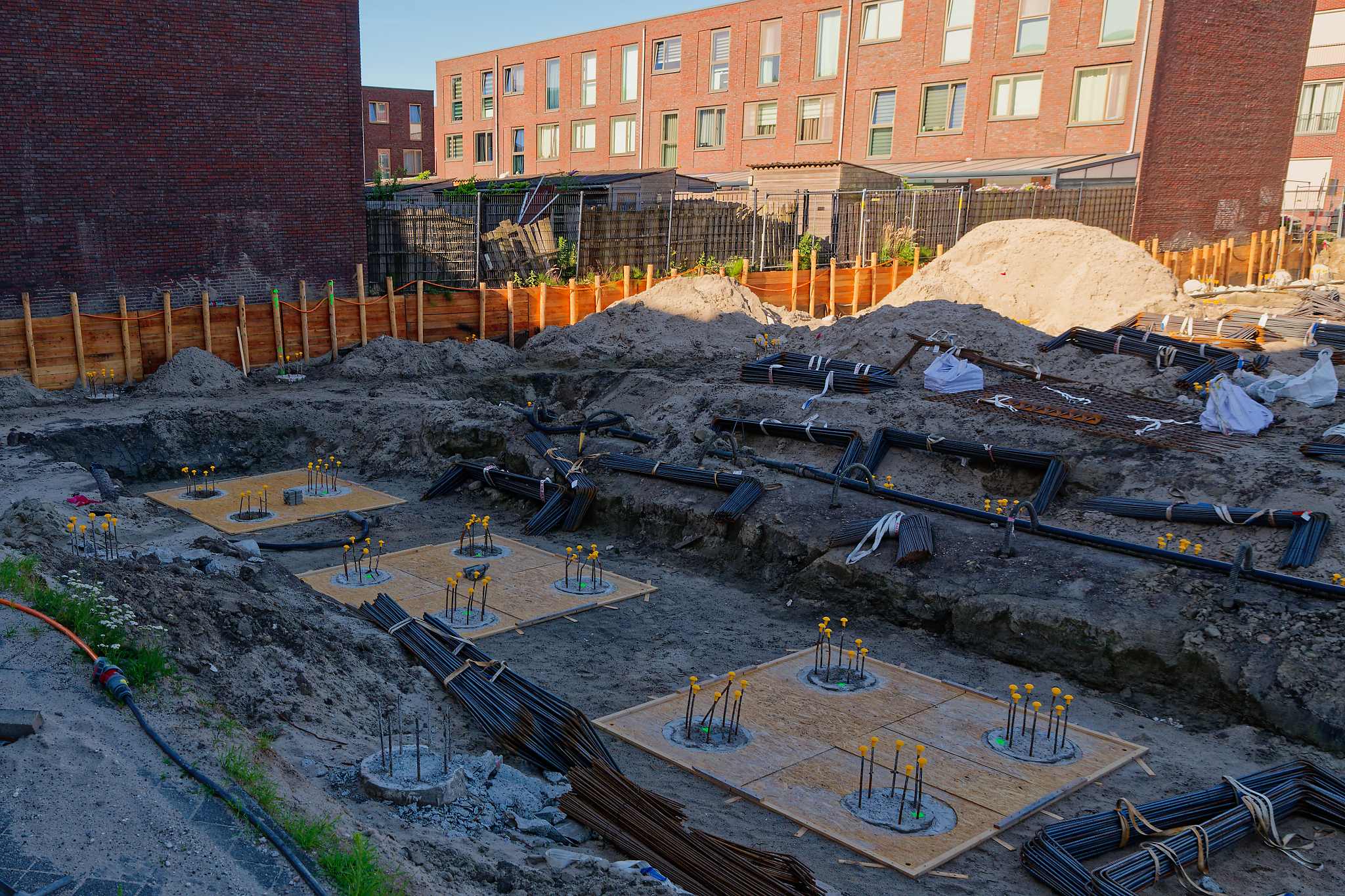Close-up view of completed pile caps in the foundation pit: square plywood bases with yellow-capped rebar stubs, black drainage membrane in the trenches between caps, bundled rebar in the foreground, orange hoarding fence and brick apartment blocks behind