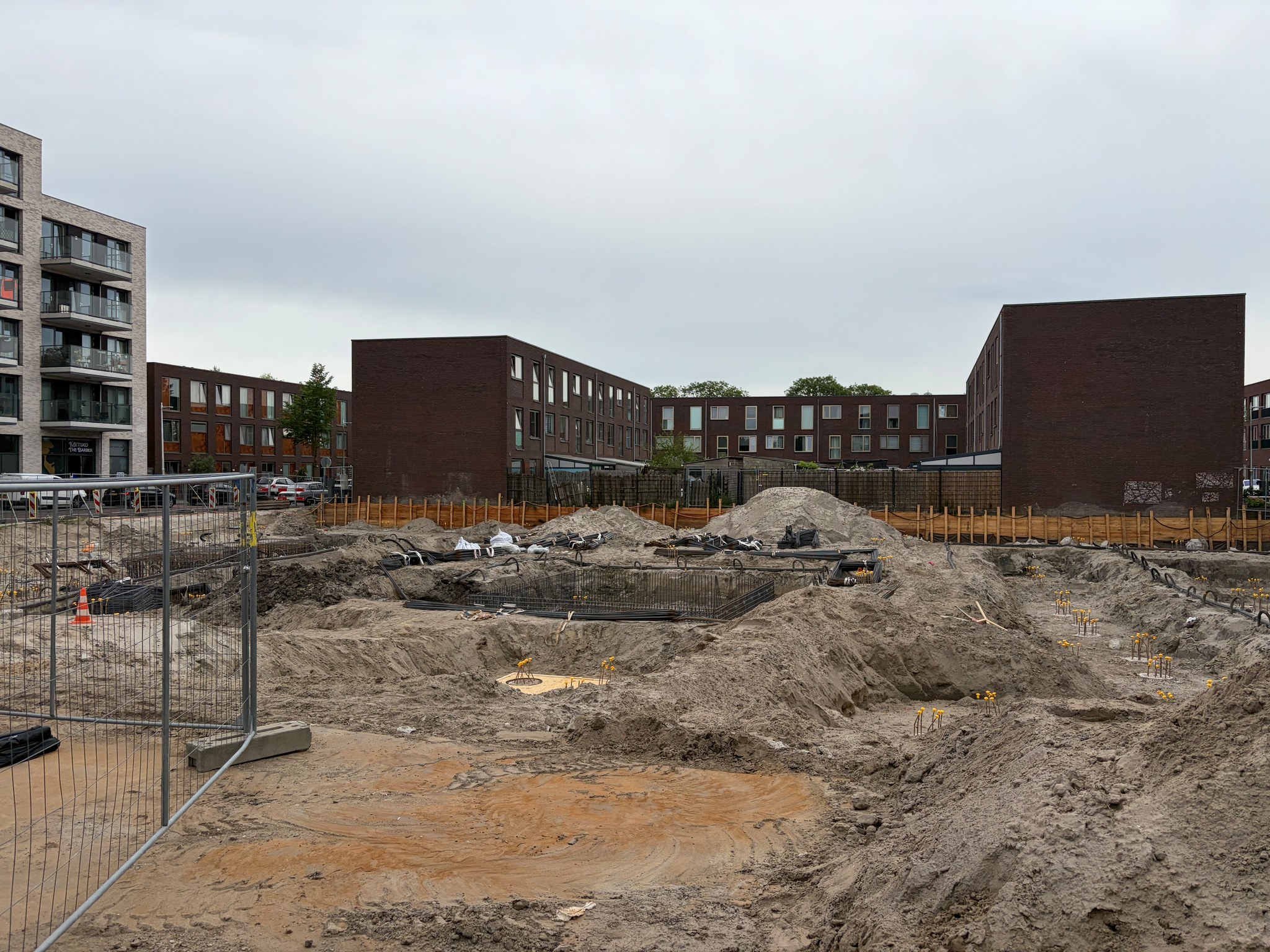 View through the street-level security fence across the Circulus foundation pit on a cloudy day: cured pile caps with yellow rebar stubs on the right, raw rebar cages assembled on sand on the left, sand mounds and timber sheet piling visible along the perimeter, neighbouring brick apartment blocks in the background
