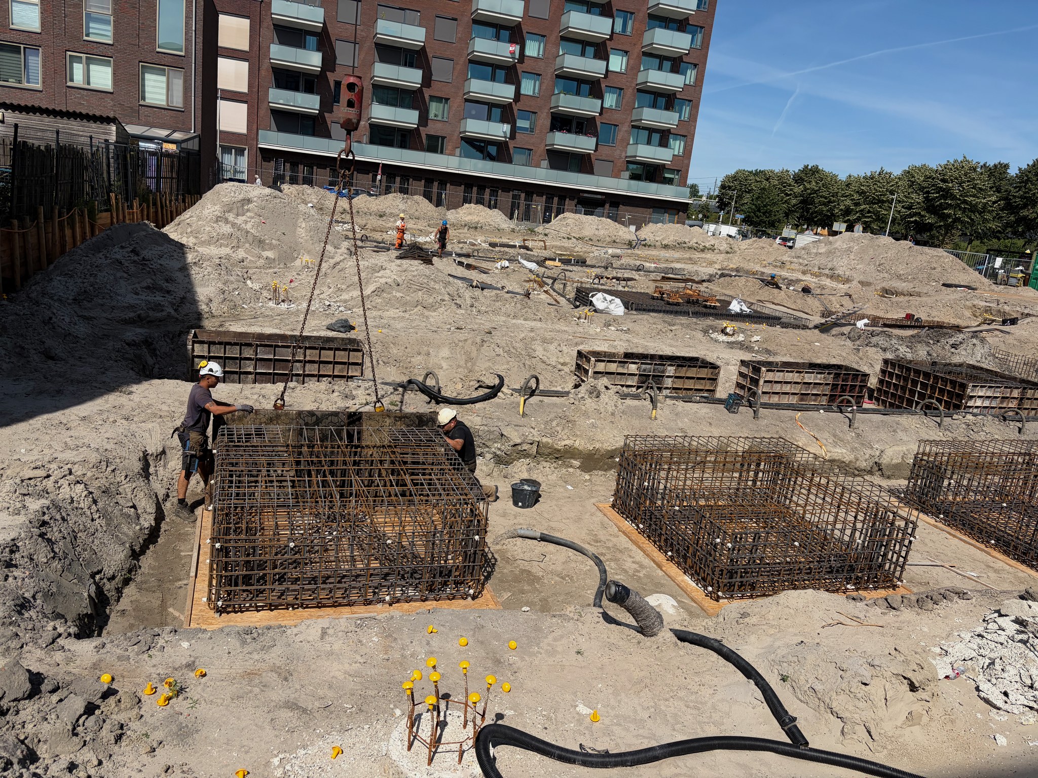 Close-up action shot at the Circulus foundation pit: two workers in hard hats guide a heavy rebar cage being lowered on a crane hook chain, plywood pile-cap base visible below, more rebar cages and sand mounds stretching into the background under a blue sky