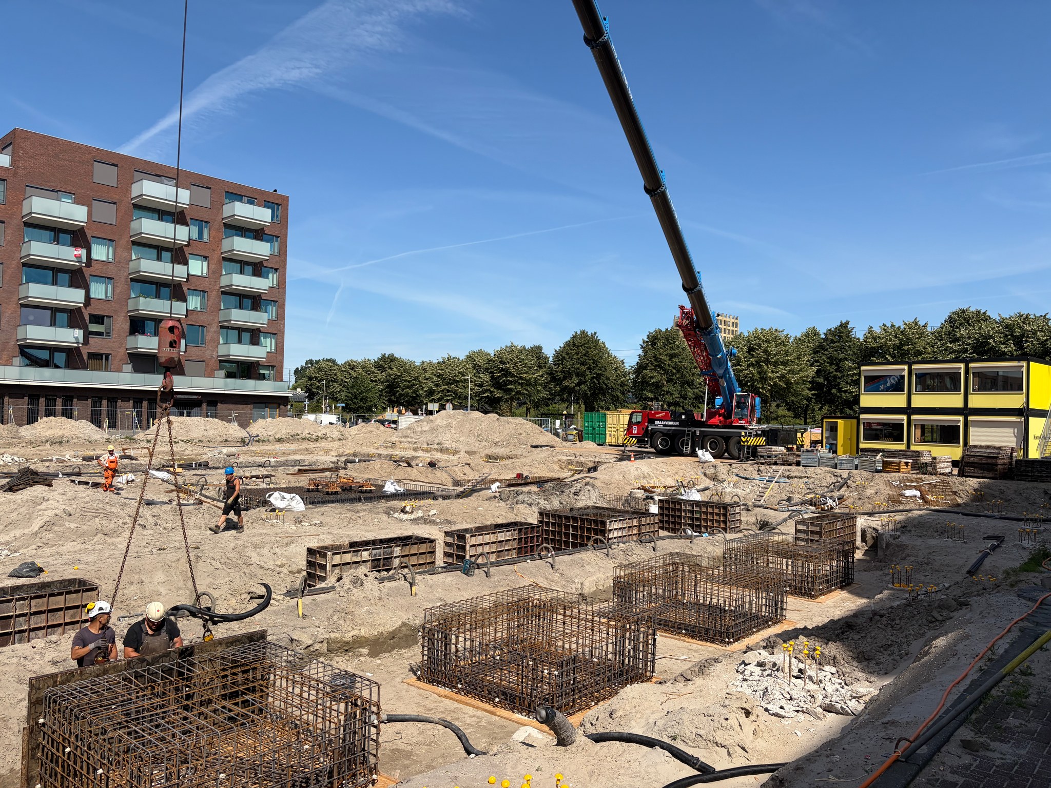 Full-site view of the Circulus foundation pit: crane boom cuts diagonally across the blue sky, workers guide a rebar cage in the foreground, rows of already-placed cages fill the centre and background, yellow site office container visible at right