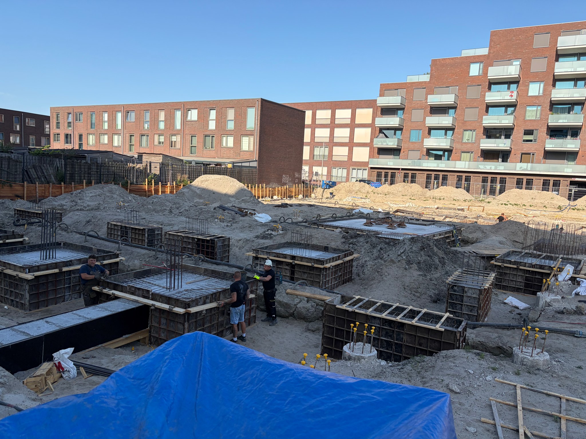 View from an elevated vantage over the Circulus foundation pit on a clear morning: rows of square steel-and-timber formwork boxes covering the rebar cages, two workers fastening panels in the centre, completed pile caps with yellow rebar visible in the right foreground, blue tarp in the lower foreground, brick apartment blocks in the background