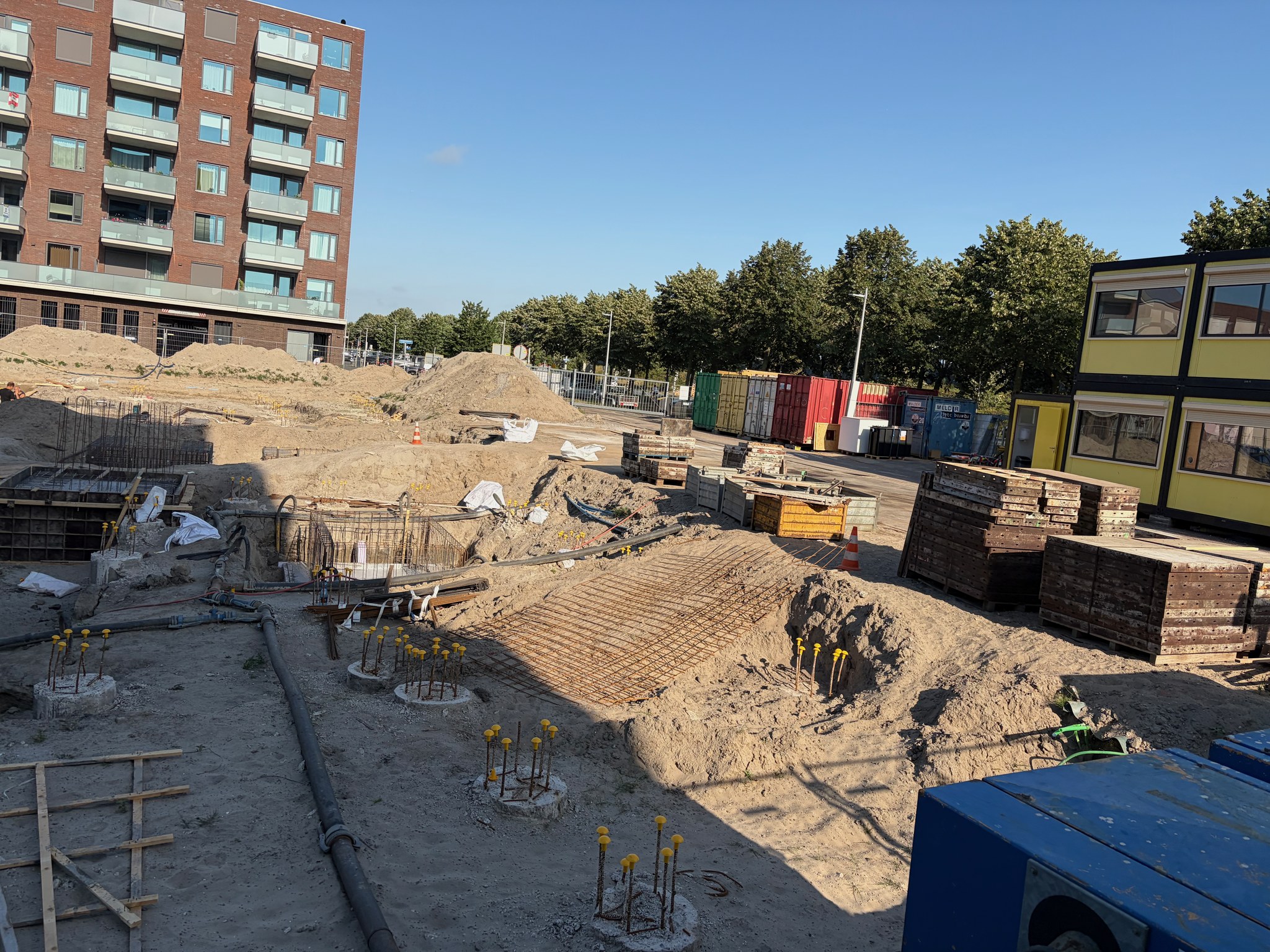 Street-level view of the Circulus foundation pit: completed stripped pile caps with yellow rebar stubs in the right foreground, a large flat rebar mat laid on sand in the centre, stacked formwork panels against the yellow site office container at right, morning sun casting long shadows across the sandy pit