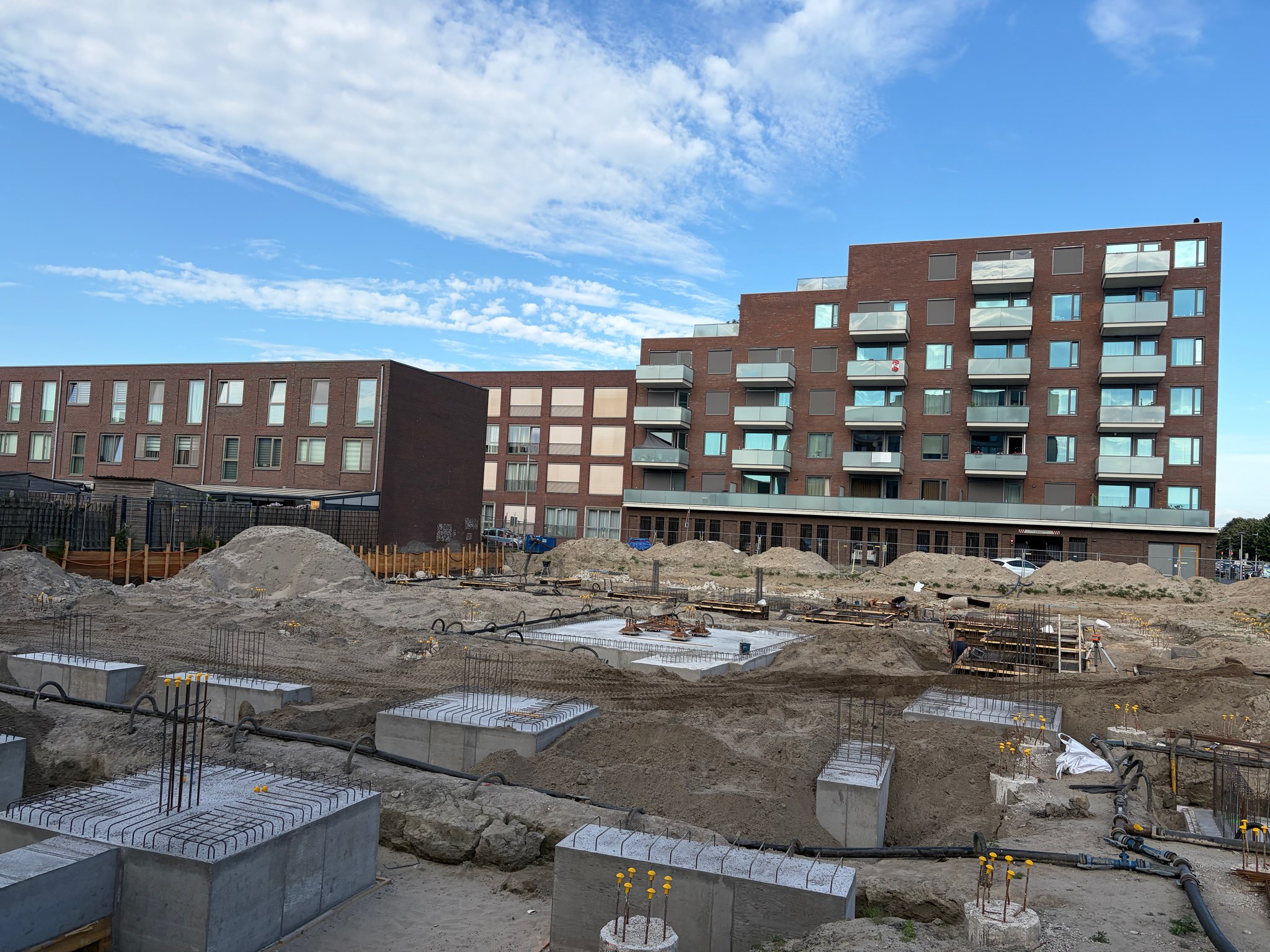 Wider view of the Circulus construction site from the street: multiple rows of stripped concrete pile caps with rebar stubs visible in the foreground, rebar grid work continuing in the middle distance, timber sheet piling along the far perimeter, brick apartment blocks behind under a partly cloudy sky