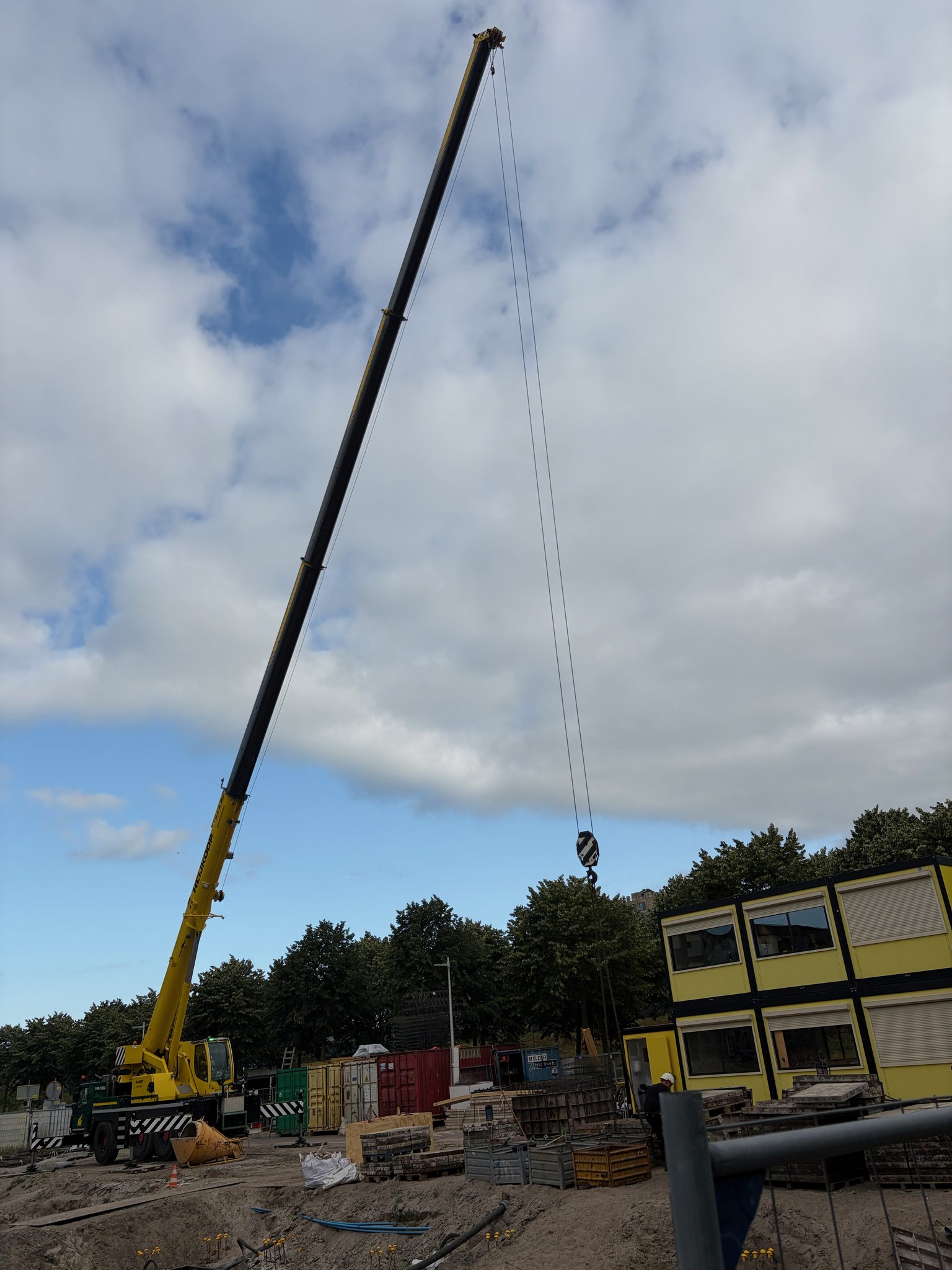 Low-angle view straight up at a yellow mobile crane with its boom raised nearly vertical against a partly cloudy sky, outriggers deployed, hook block hanging free, shipping containers and a two-storey yellow site office visible at the base