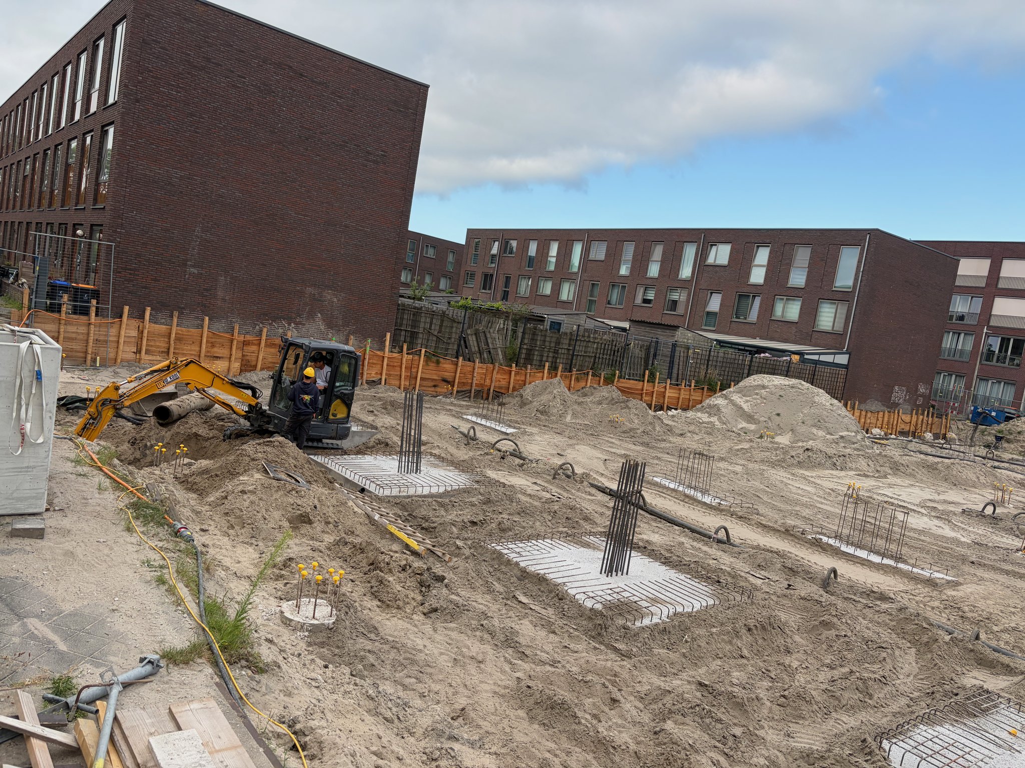 Mini excavator with two workers laying flat rebar ground-beam mats against a timber-faced retaining wall at the rear boundary of the Circulus site, rows of pile caps with vertical rebar stubs in the foreground, brick apartment buildings visible behind the fence line