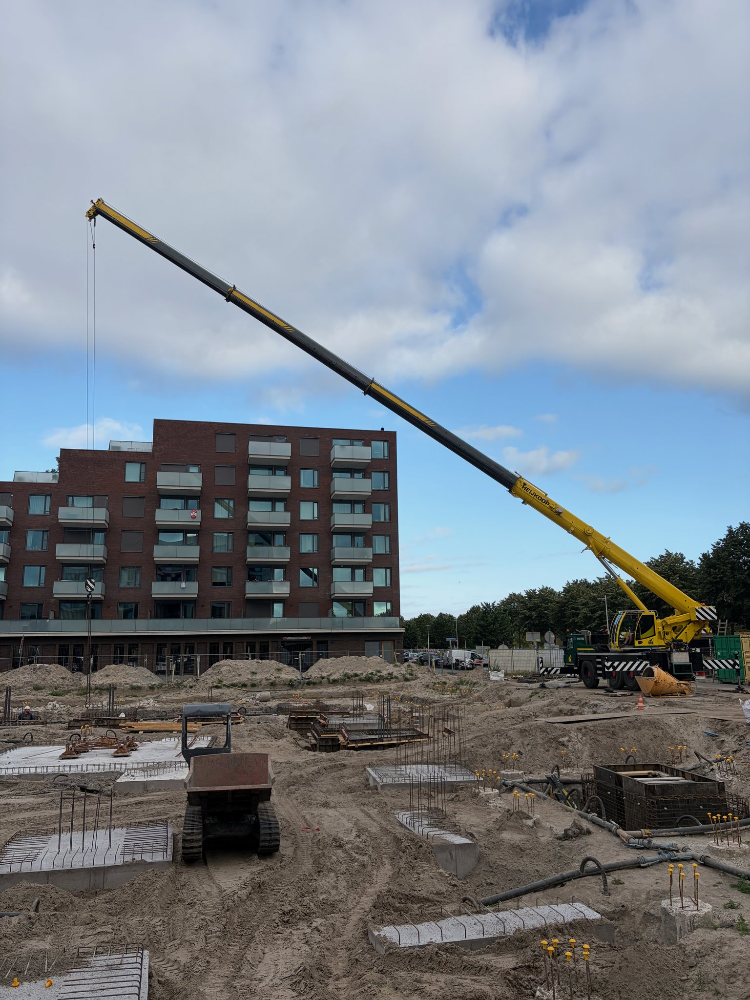 Yellow mobile crane with Heijkoop branding on its boom, fully extended low over the Circulus construction site toward a brick apartment building in the background, foundation pit with rebar-topped pile caps and a tracked dumper in the foreground