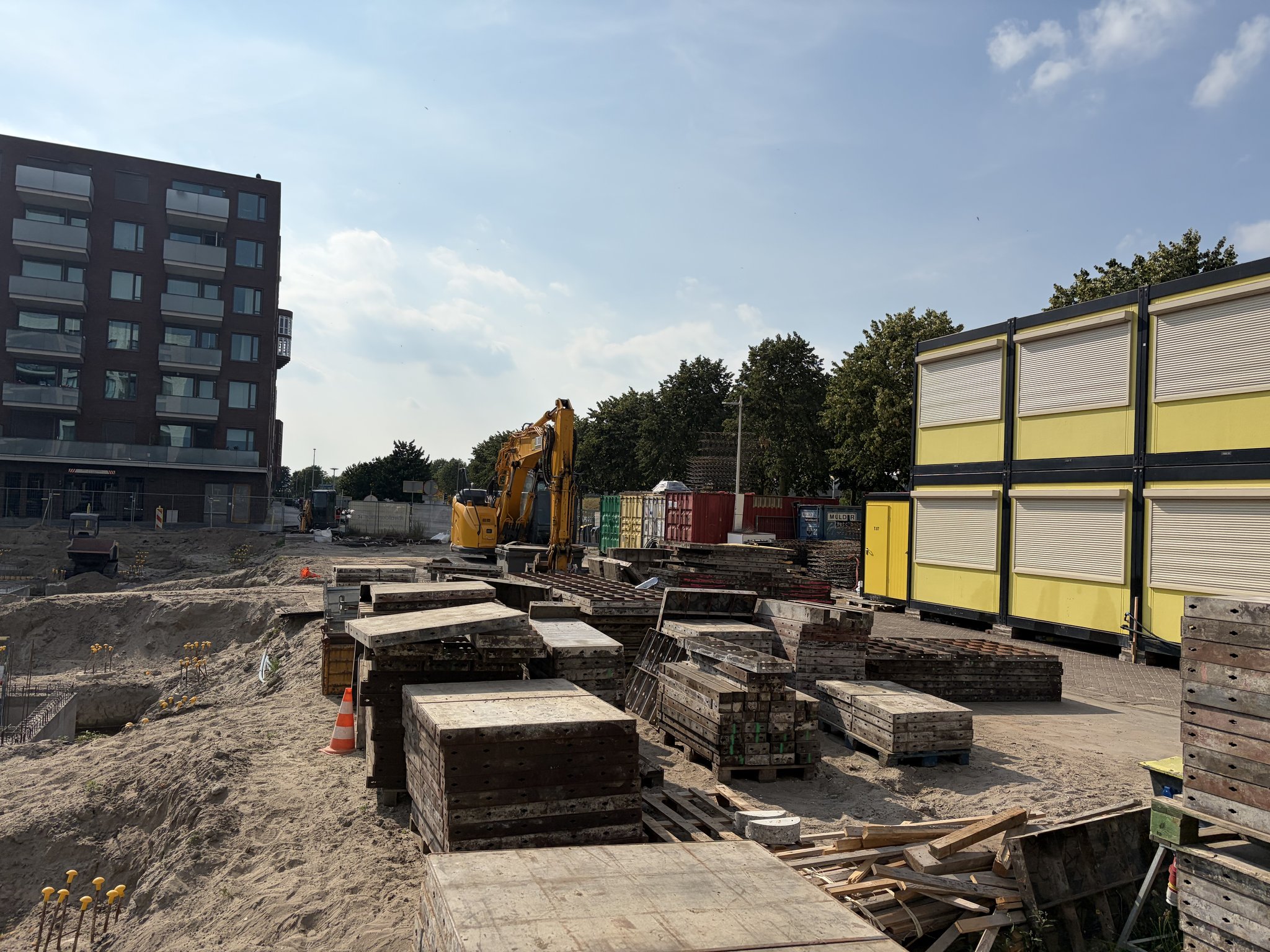 Stacks of used timber-and-steel formwork panels and loose timber on sand beside the Circulus site entrance, a yellow excavator and double-stacked yellow site cabins behind, a brick apartment block with balconies on the left under a summer afternoon sky