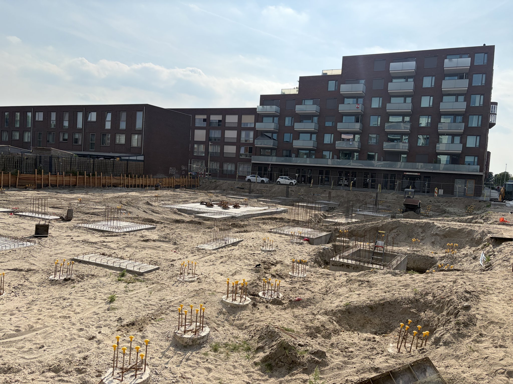 The Circulus site in late-afternoon sun: rebar-topped pile caps with yellow safety caps across the sand, flat rebar mats and a wide square concrete pad with steel anchors in the middle of the plot, a timber retaining wall and brick apartment blocks with balconies at the back