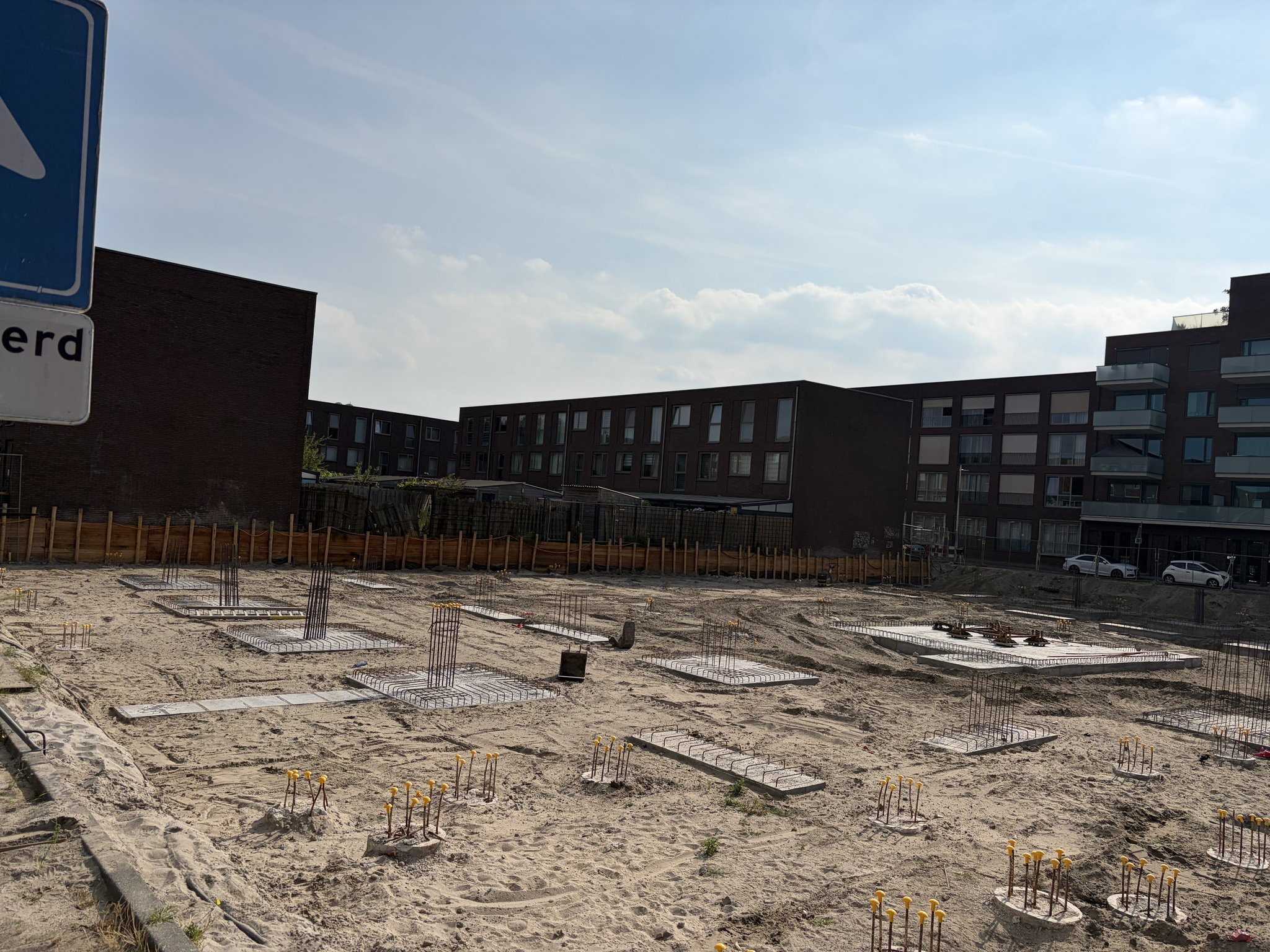 View from the street corner past a blue road sign across the Circulus site: pile caps with yellow-capped rebar starters, flat rebar mats and a square concrete pad on sand, timber retaining wall and dark brick terraced houses and apartment blocks behind