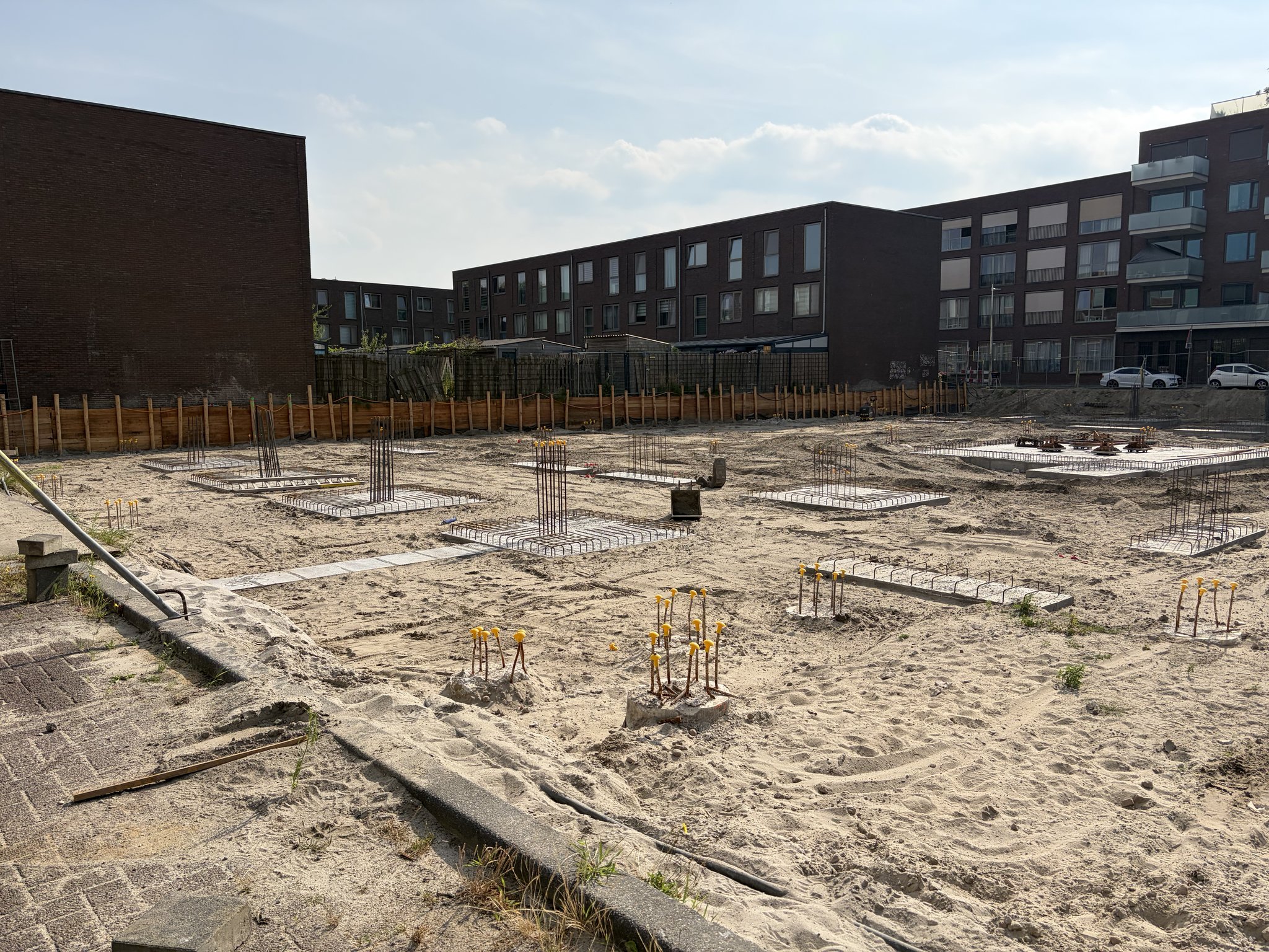Looking along the edge of the Circulus foundation pit from a paved kerb: pile caps with yellow-capped starter bars in the foreground, rebar mats and cages further back, the orange timber retaining wall along the boundary and brick apartment buildings behind