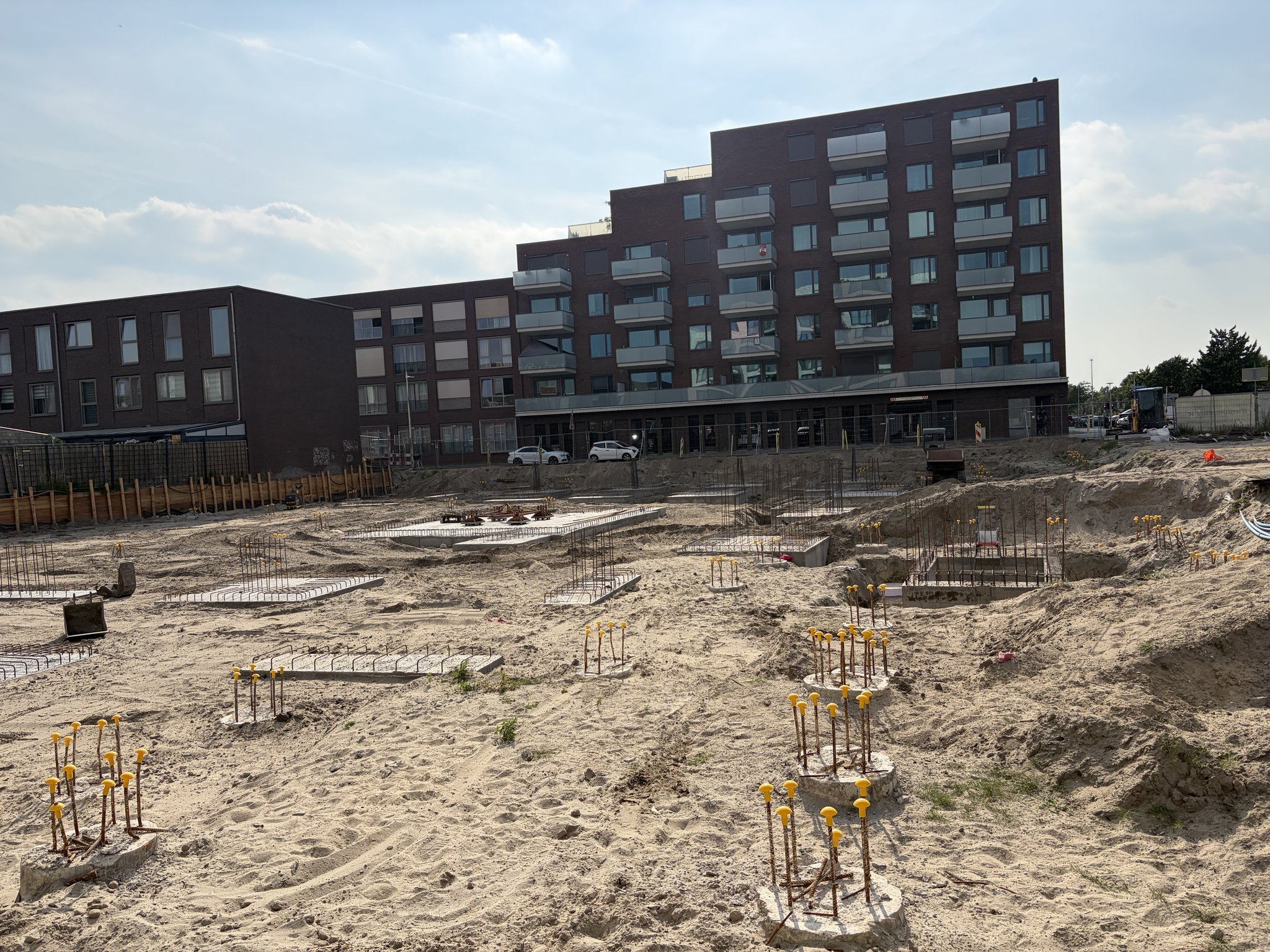 Late-afternoon overview of the Circulus site: clusters of rebar starters with yellow caps on low concrete pile caps across pale sand, flat rebar mats, a square concrete pad and a mound of sand on the right, a seven-storey brick apartment block with glass balconies in the background