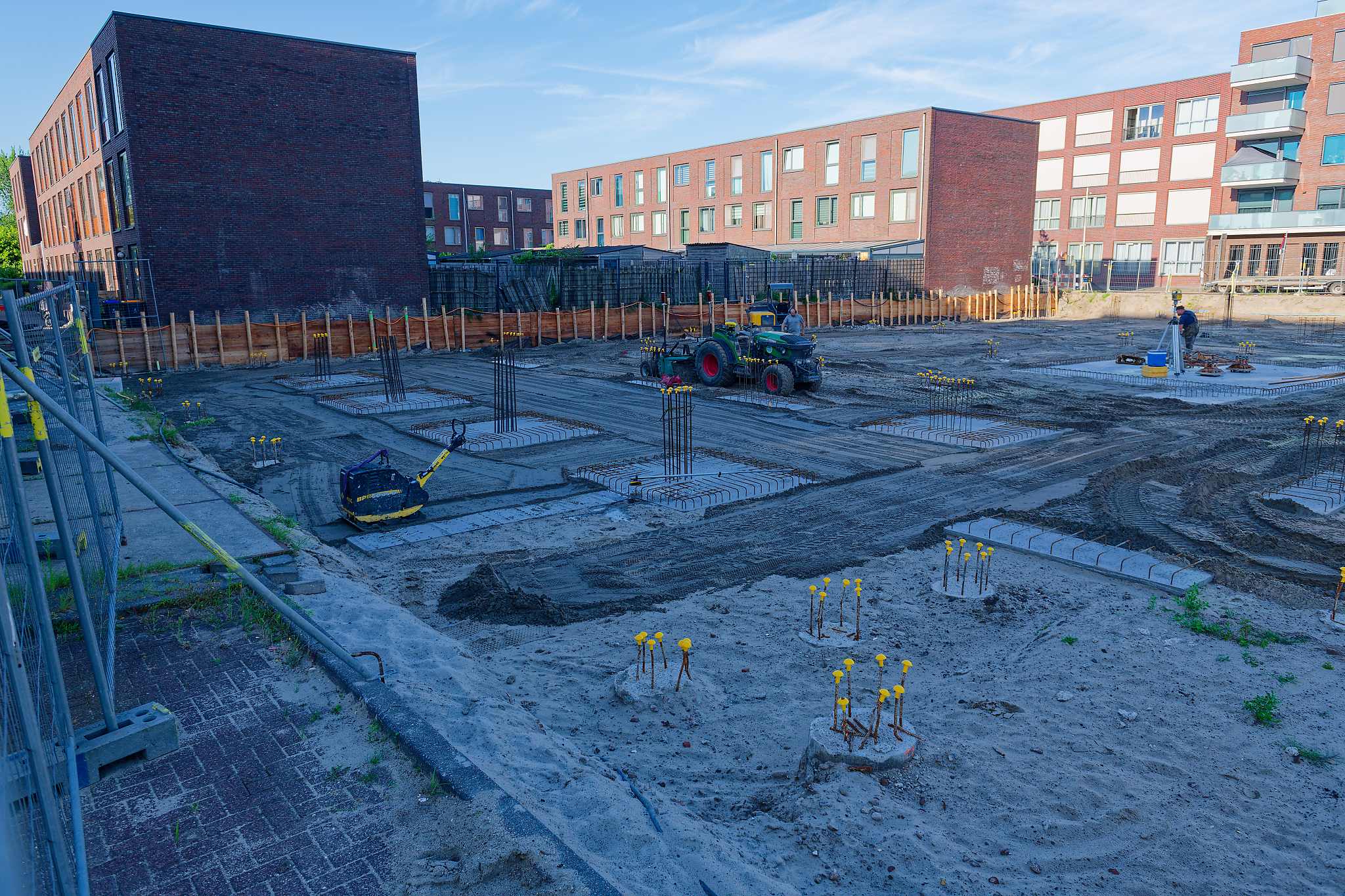 Construction site with a yellow plate compactor parked among rows of square rebar cages on concrete pads, a John Deere tractor and surveyor with tripod further back, wooden retaining wall and dark brick building along the boundary, morning light