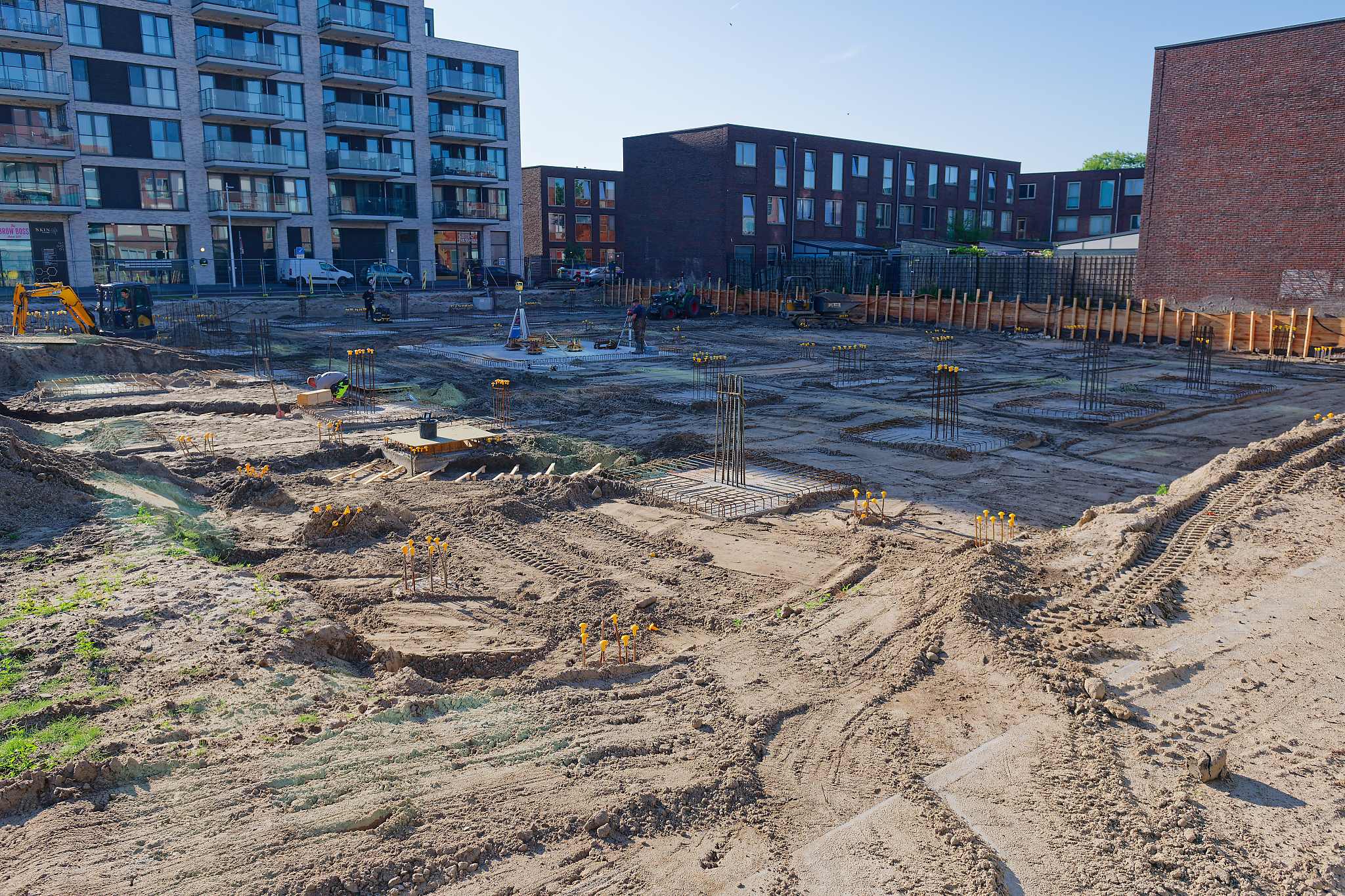 Panoramic view of the Circulus site from the street side, showing rows of rebar-topped pile caps spreading across sandy ground toward a dark brick apartment building, a mini excavator and tractor at work, taller grey apartment blocks with balconies on the left under a clear morning sky