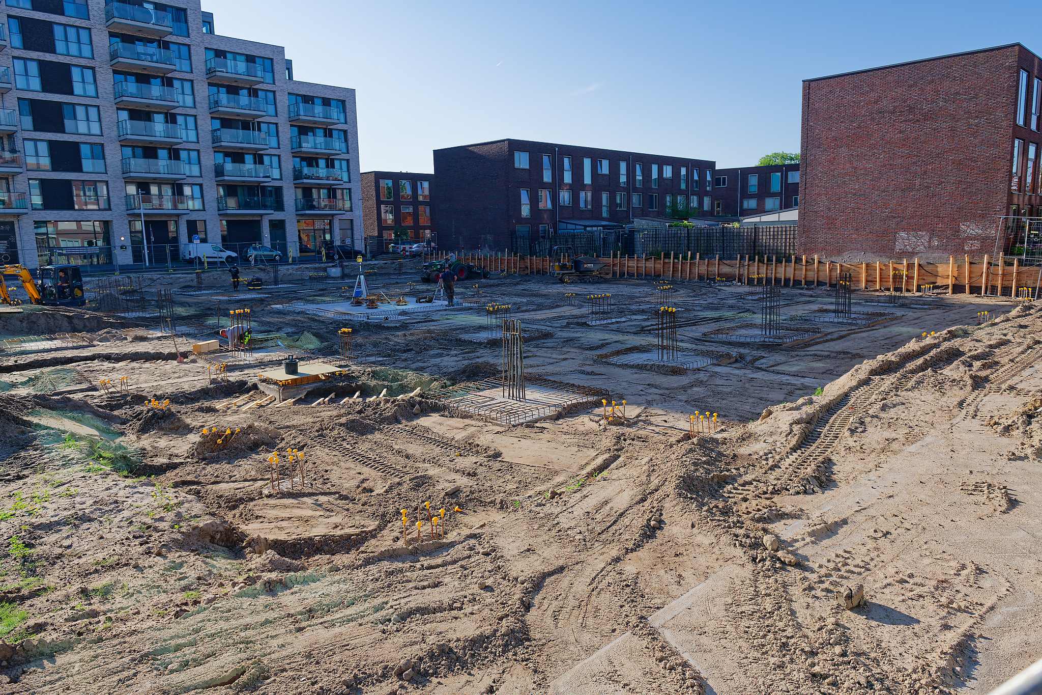 Elevated view across the Circulus foundation grid toward apartment buildings, showing rows of pile caps with vertical rebar, a formwork box under construction in the foreground, and a steep embankment of excavated sand along the right side of the site
