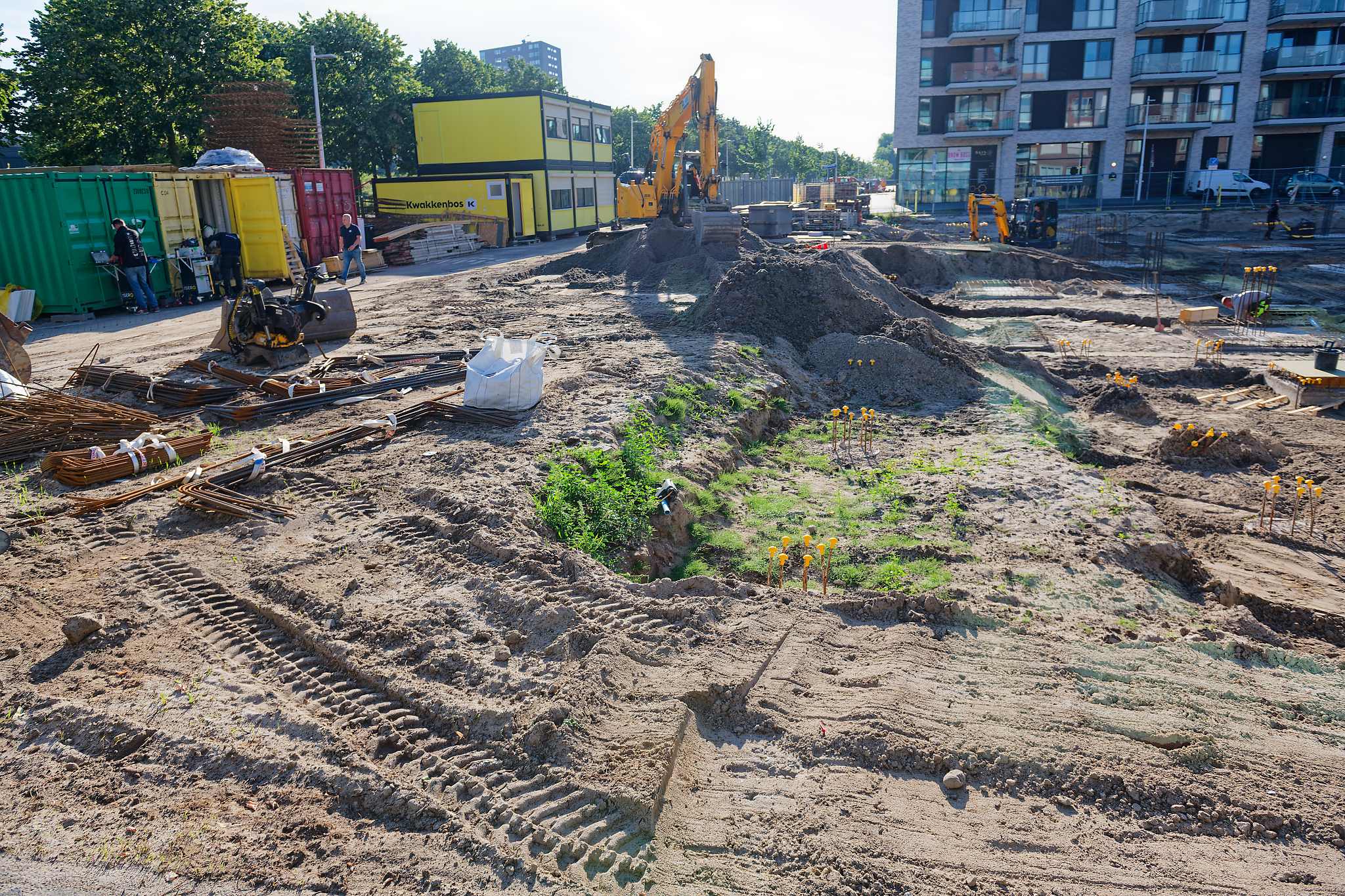 Site entrance area with stacked green, red and white shipping containers, a yellow Kwakkenbos site cabin, bundled rebar and coiled orange conduit on the ground, a yellow excavator digging a long trench past a weedy older cut, apartment buildings in the background