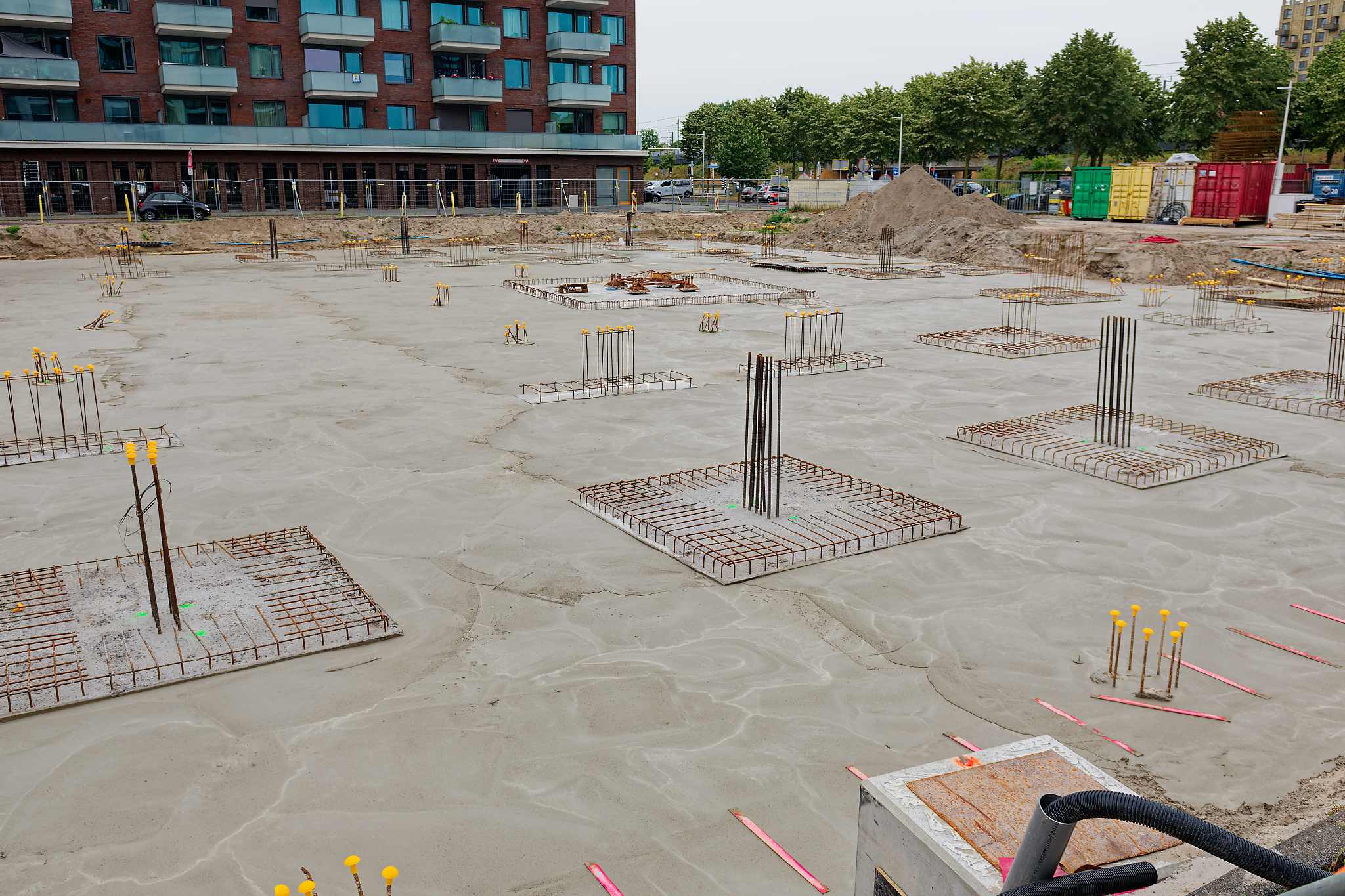 Wide panoramic view of the Circulus site from a raised vantage, the poured concrete floor slab spreading across the foreground with rows of yellow-capped rebar starters, stacked green, yellow and red shipping containers and a mound of excavated sand at the site edge, tree-lined street and apartment blocks in the background
