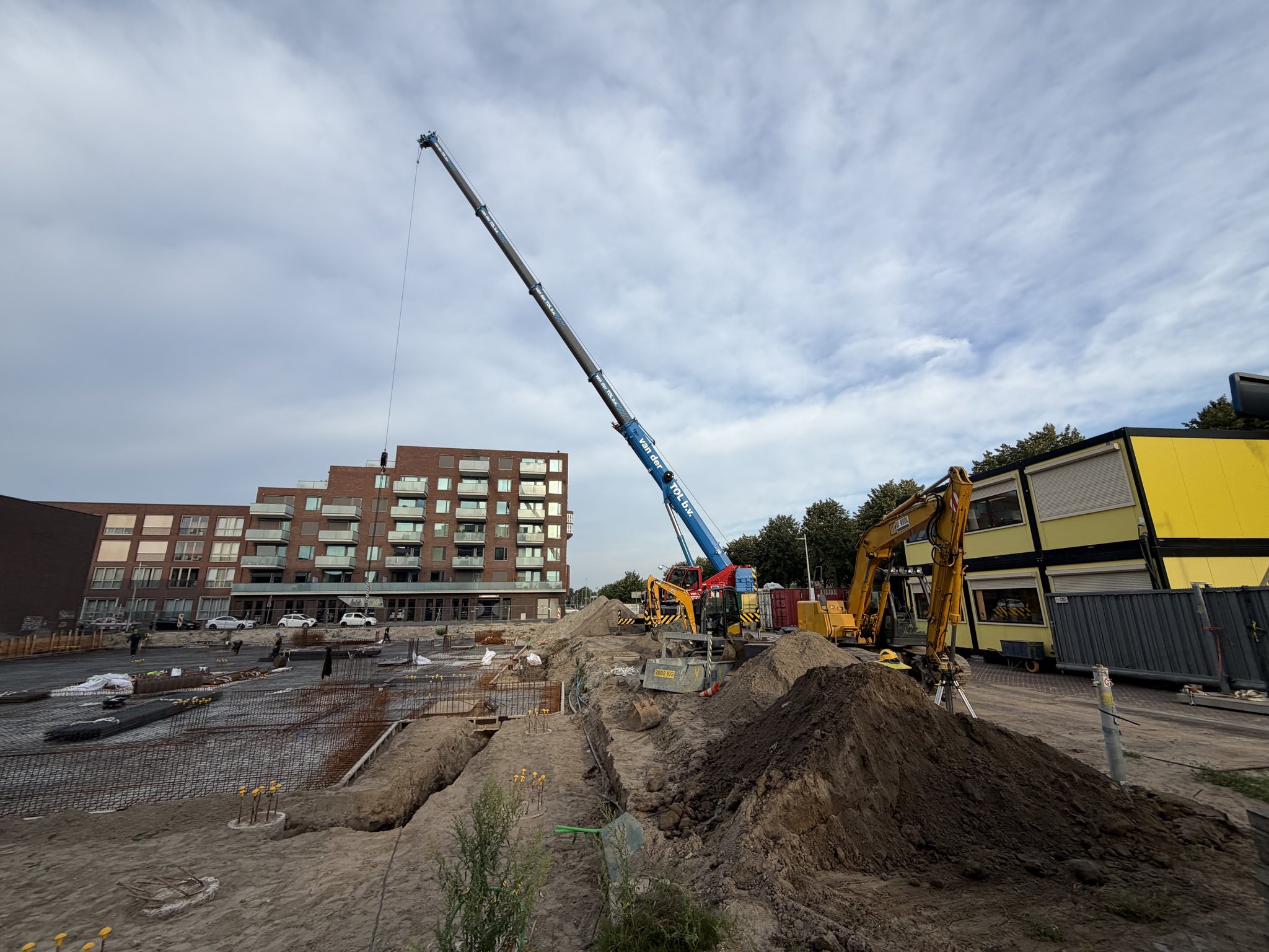 A blue van der Tol mobile crane with its boom raised high over the Circulus site entrance, a yellow excavator beside a large heap of dug soil and a long service trench, rebar mesh and formwork edges in the foreground, yellow site cabins and brick apartment blocks under a cloudy sky