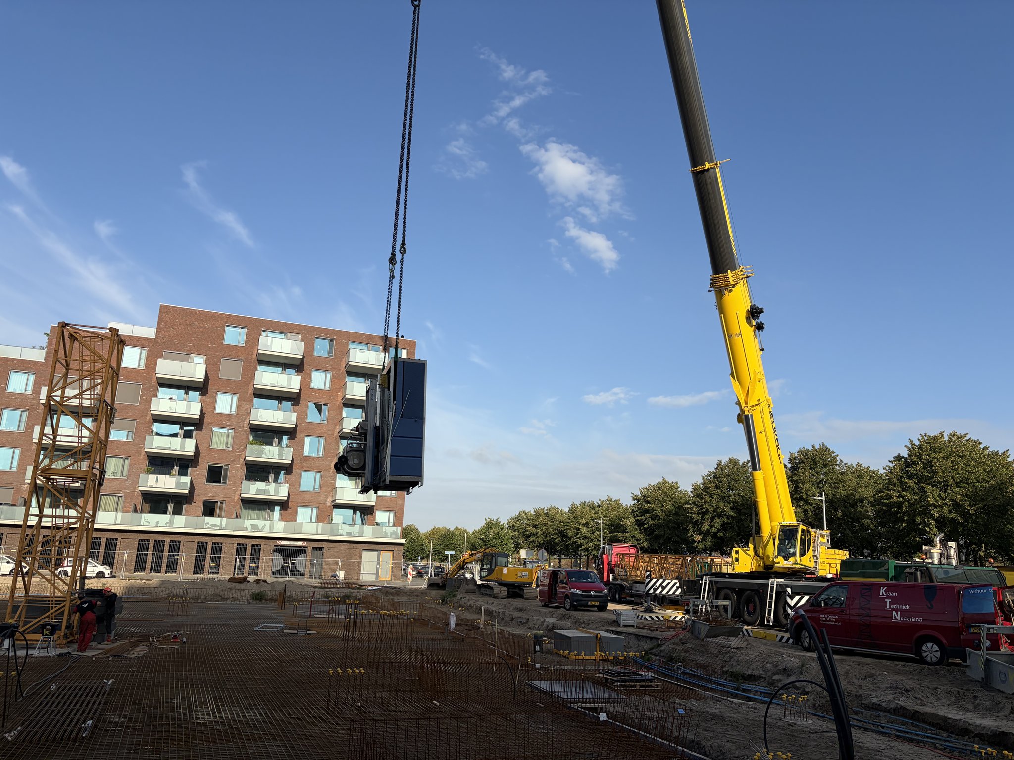A yellow mobile crane lifting a dark blue tower-crane machinery unit on chains against a clear blue sky, the first orange lattice mast sections of a tower crane standing on the left with fitters at the base, a rebar mesh deck, excavator, red Kraan Techniek Nederland vans and a lorry with mast sections on the right