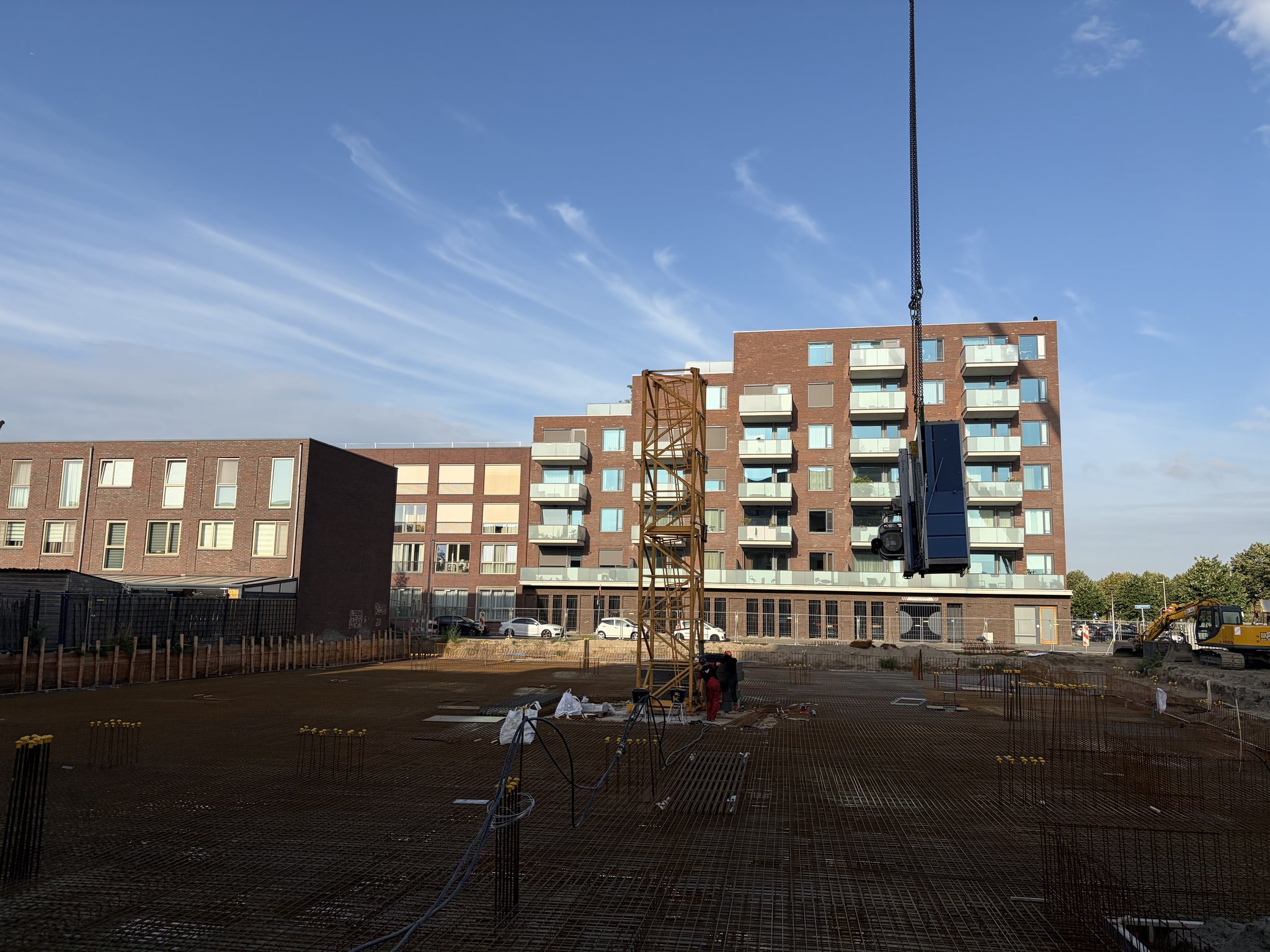 The dark machinery unit hanging from the mobile crane's hook beside the growing orange lattice tower crane mast on the Circulus site, rebar mesh deck and black cable ducts in the foreground, brick apartment building with balconies behind under a blue sky