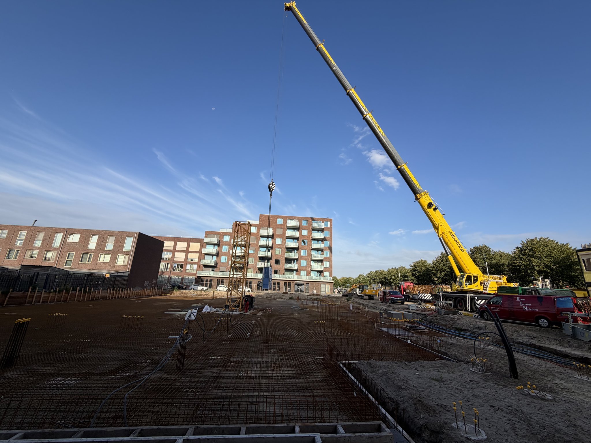 Wide-angle view across the rust-brown rebar mesh deck of the Circulus site toward a yellow mobile crane with its boom extended high over a partly built orange lattice tower crane mast, red vans parked beside it, brick apartment blocks under a deep blue morning sky