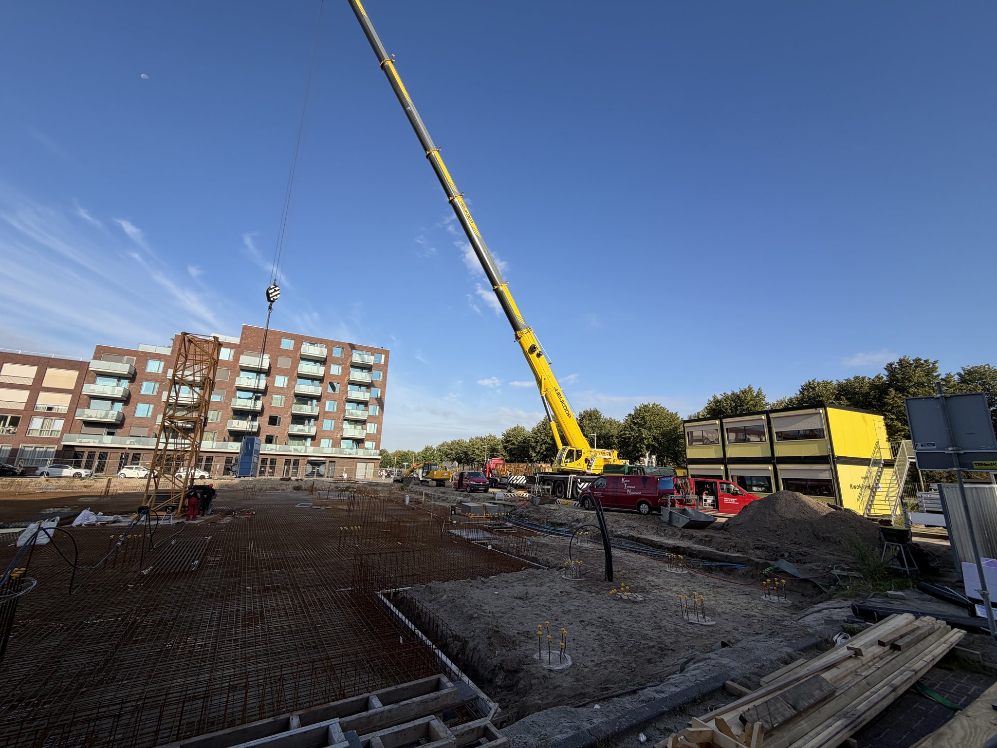 The Circulus site from the entrance during tower crane assembly: yellow mobile crane boom raised, the lattice mast on its pad in the middle of the rebar deck, stacked yellow site cabins, a sand heap and timber formwork boards in the foreground