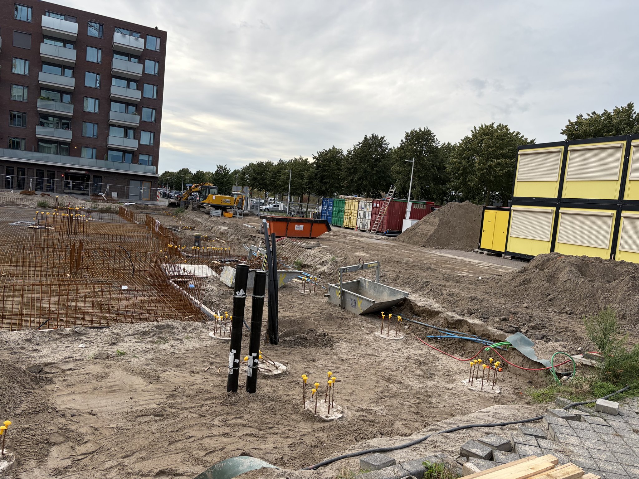 Entrance corner of the Circulus site: two black utility ducts standing upright in the sand beside yellow-capped pile caps, a trench with red, blue and green conduit, a galvanised concrete skip, rebar cages on the left, an orange skip, coloured containers, a sand heap and yellow site cabins