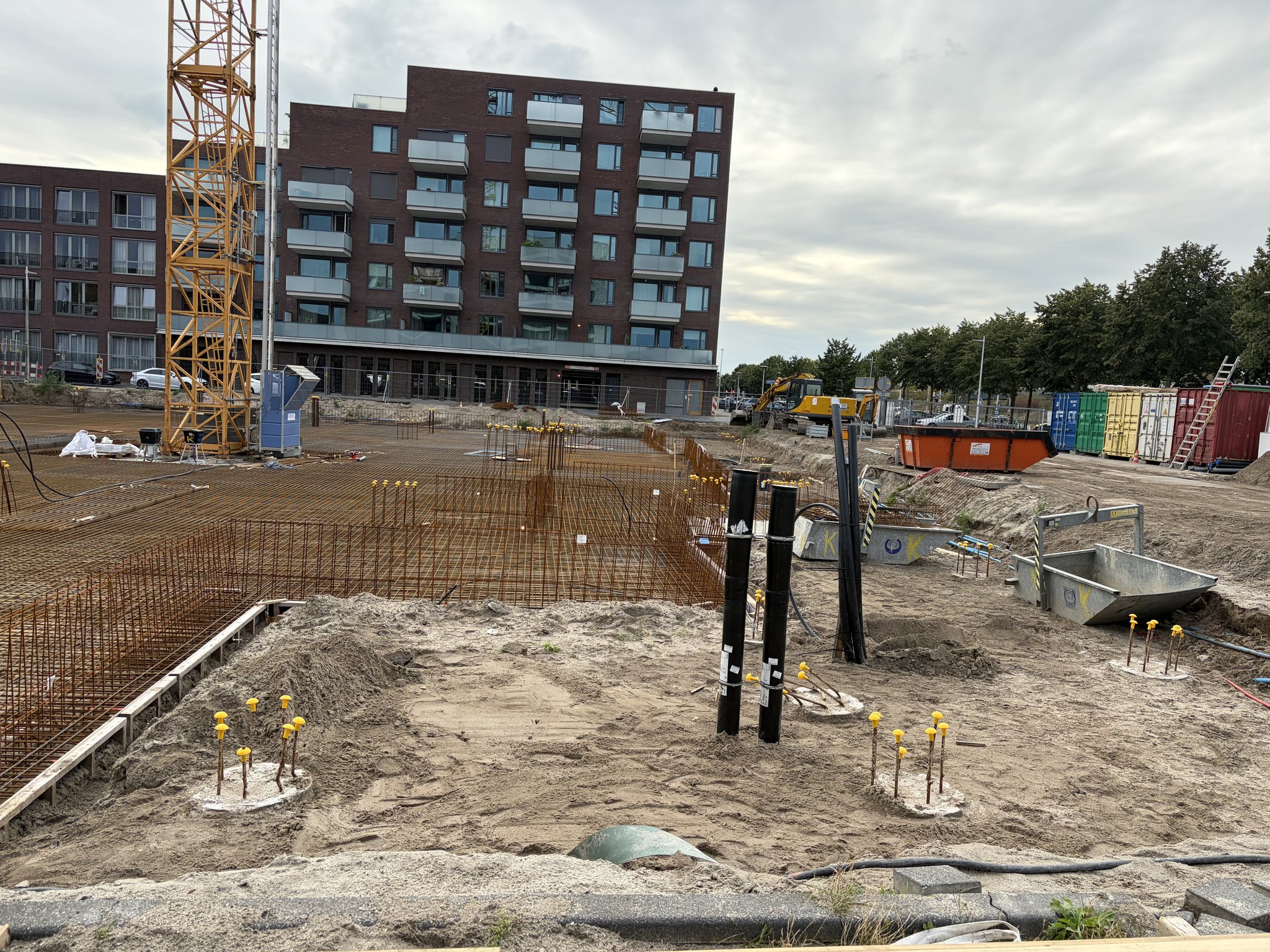 Black upright utility ducts and yellow-capped starter bars in the sand in front of the rebar mesh deck, the base of the new yellow tower crane mast and a seven-storey brick apartment block behind, coloured containers along the site edge