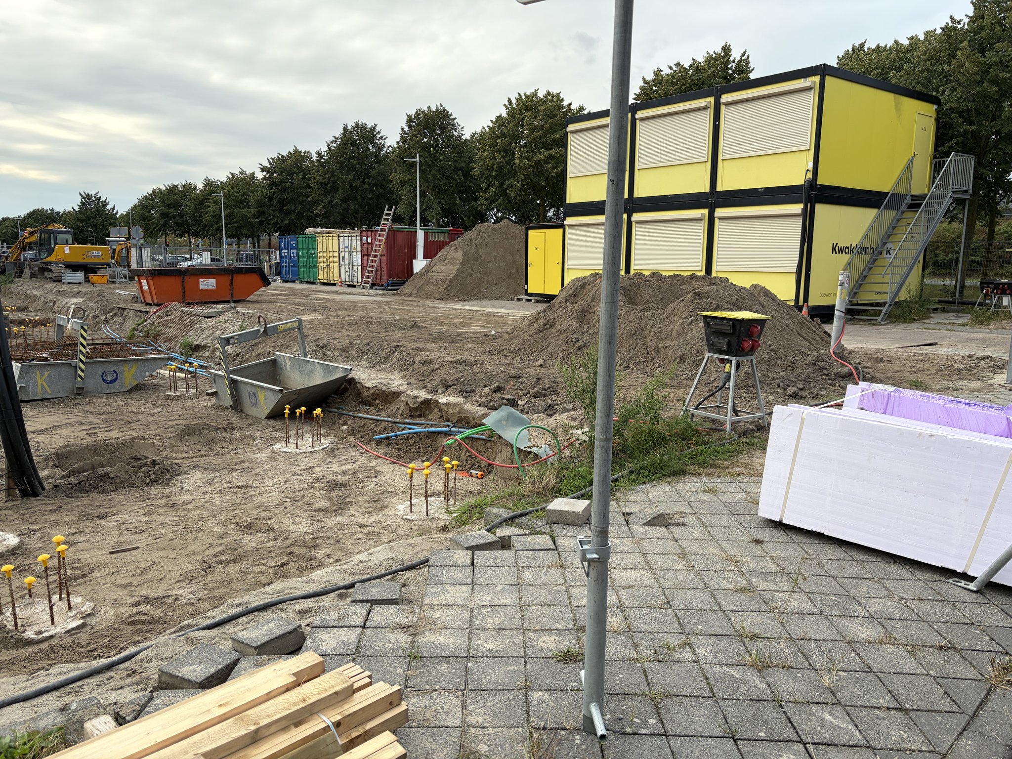Double-stacked yellow Kwakkenbos site cabins behind a heap of sand, a pallet of white and pink insulation boards and a red-topped mortar tub beside a scaffold pole in the foreground, a service trench with coloured conduit and yellow-capped pile caps on the left
