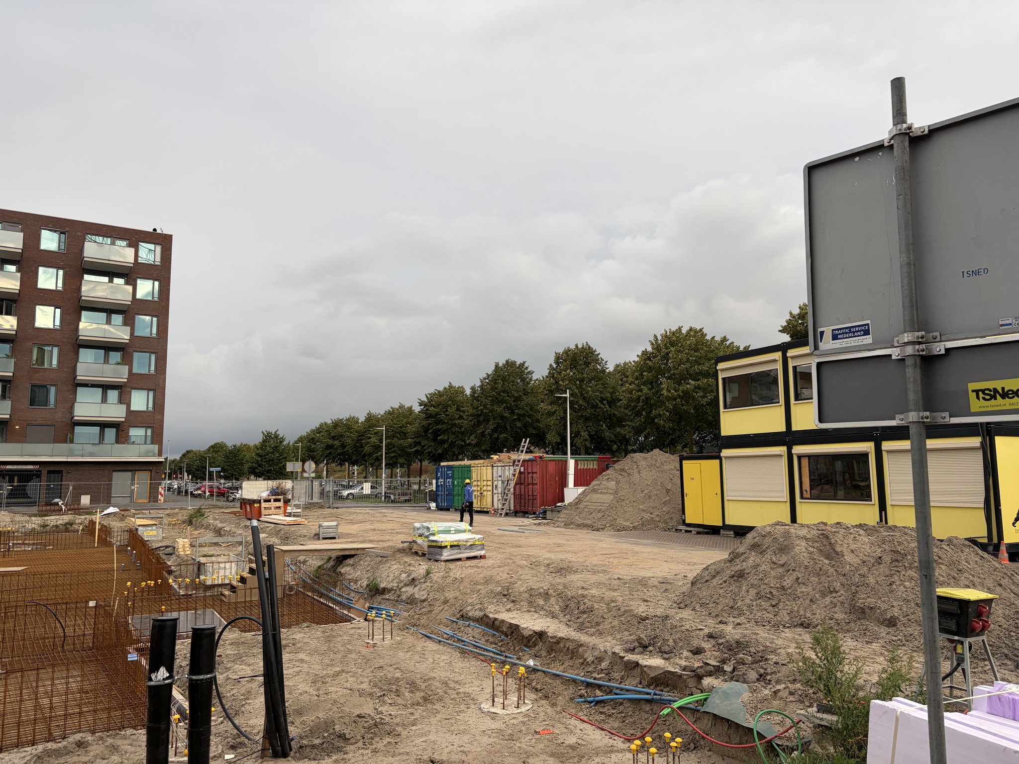 View past a road sign toward the Circulus site entrance: a service trench with blue conduit, black utility ducts, yellow-capped pile caps and a rebar cage on the left, sand heaps, coloured containers and yellow site cabins behind, pink insulation boards in the corner