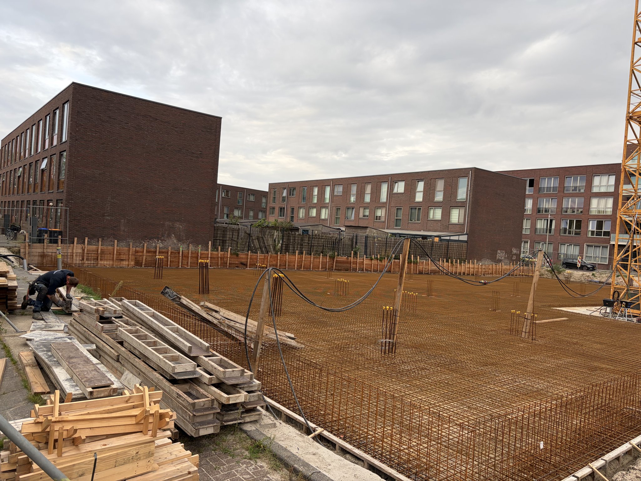 A worker kneeling on the pavement cutting timber beside stacks of used formwork boards and wooden stakes, the finished rust-brown rebar mesh deck with column starter bars stretching toward the timber retaining wall and brick terraced houses, cables slung on timber posts, tower crane mast at the right edge