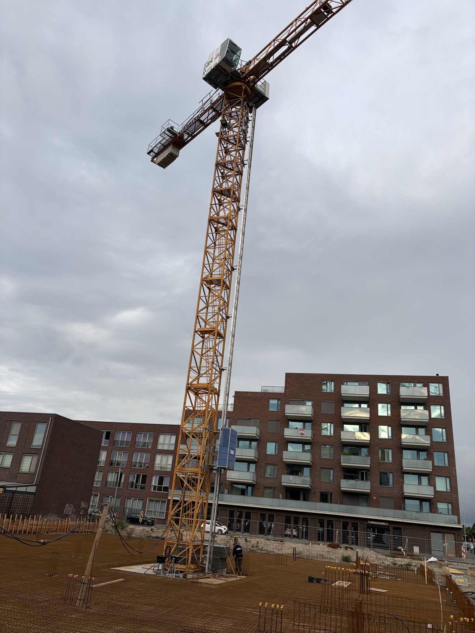 Looking straight up the yellow lattice mast of the Circulus tower crane to its cab, slewing ring and jib against a grey overcast sky, a blue electrical cabinet partway up the mast and the top floors of a brick apartment block at the bottom