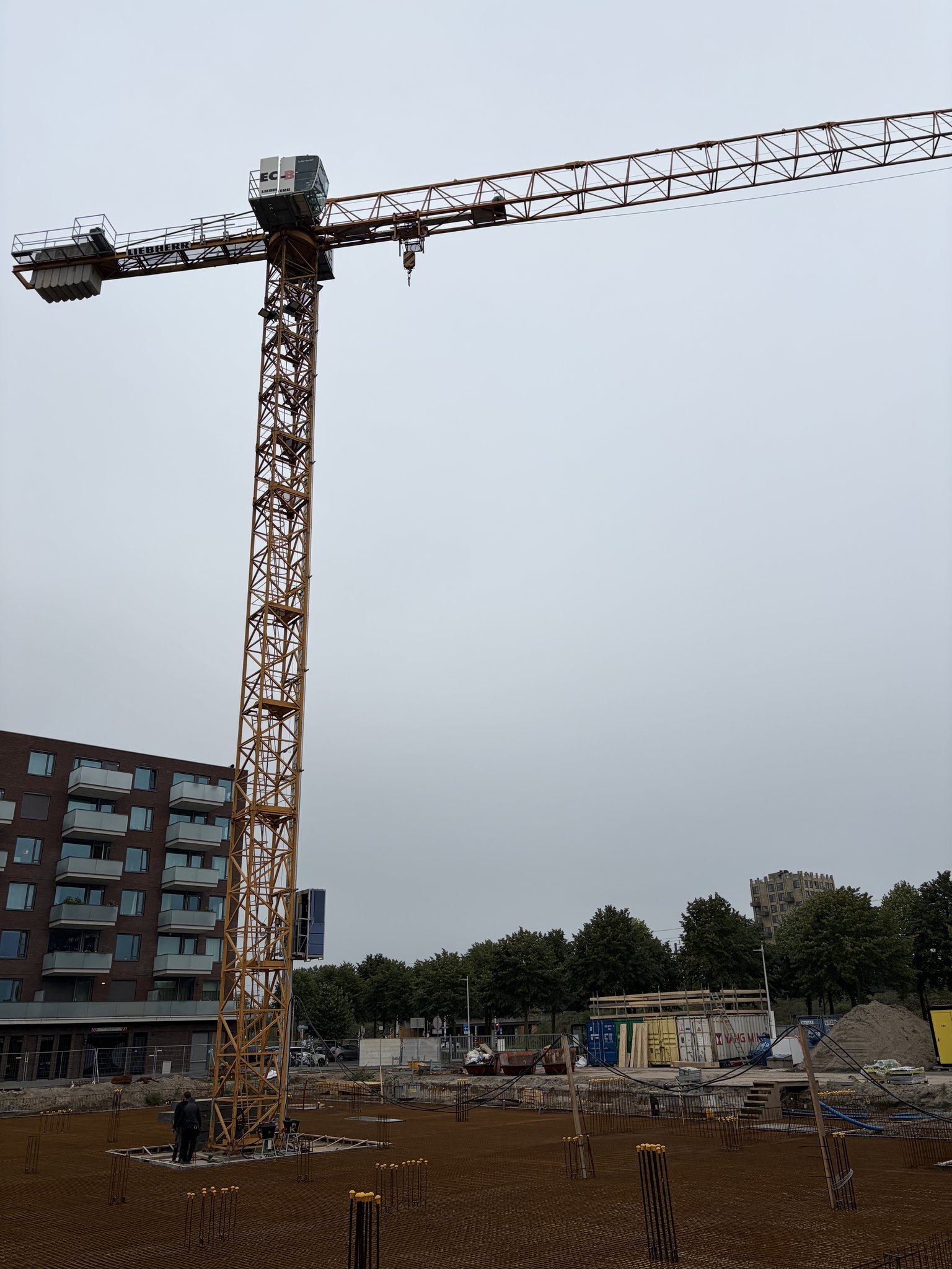 The Circulus tower crane seen from below against a pale grey sky: yellow lattice mast, operator cab and slewing unit, long horizontal jib reaching right and counter-jib with ballast to the left, treetops and a brick apartment block at the bottom