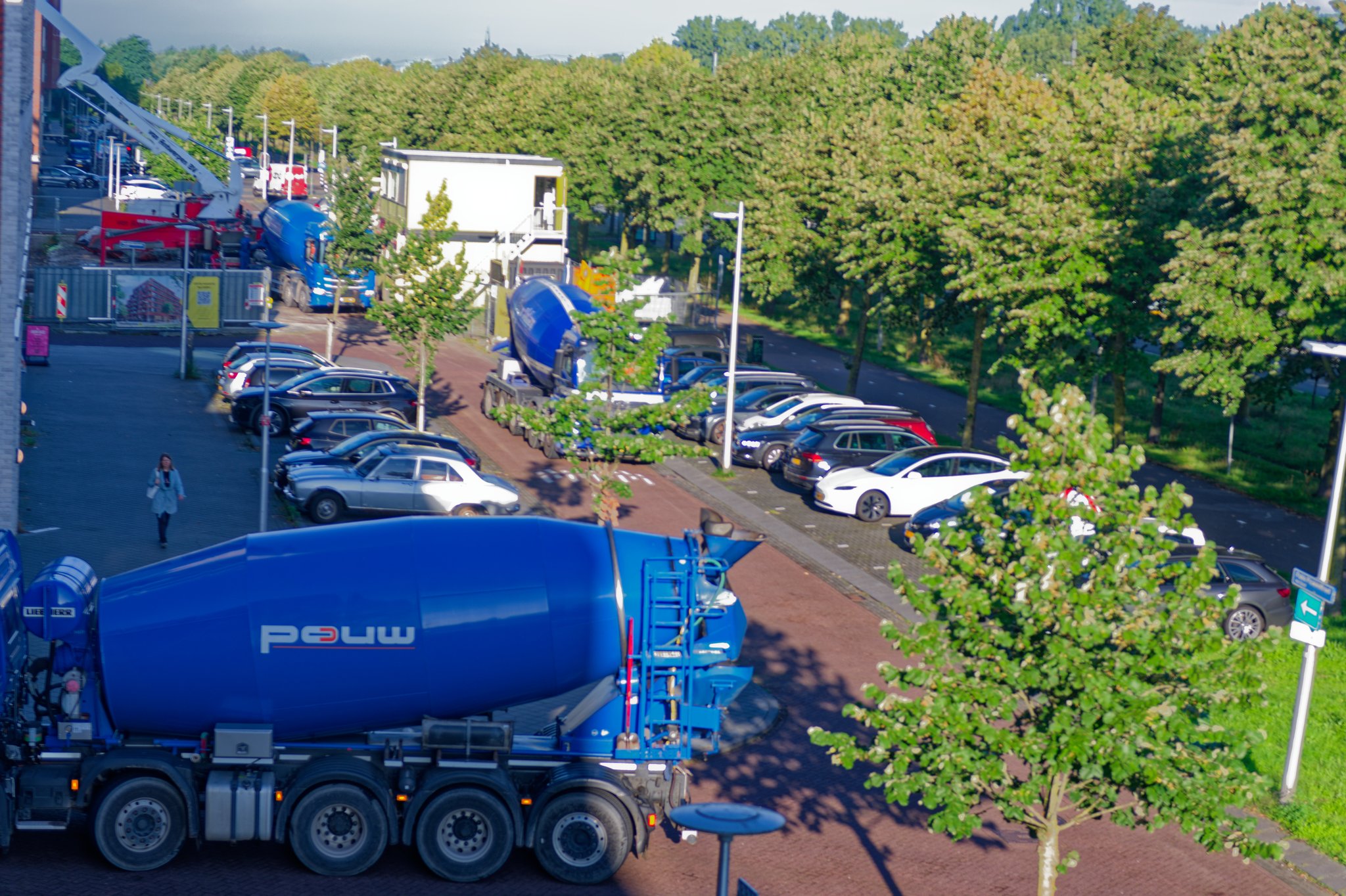 View from an upper window of a tree-lined street in Utrecht in morning sun: a blue Pouw concrete mixer truck in the foreground, more blue mixers queued past rows of parked cars, and a red truck-mounted concrete pump with its white boom unfolded beside the Circulus site hoarding and a white site office